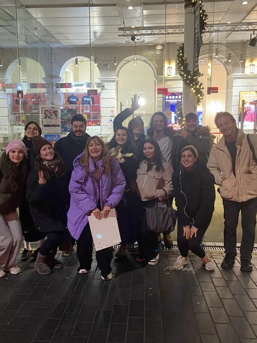 A group of people posing outside at night in front of a storefront decorated with holiday lights and garland. The group is smiling and dressed warmly for cold weather.