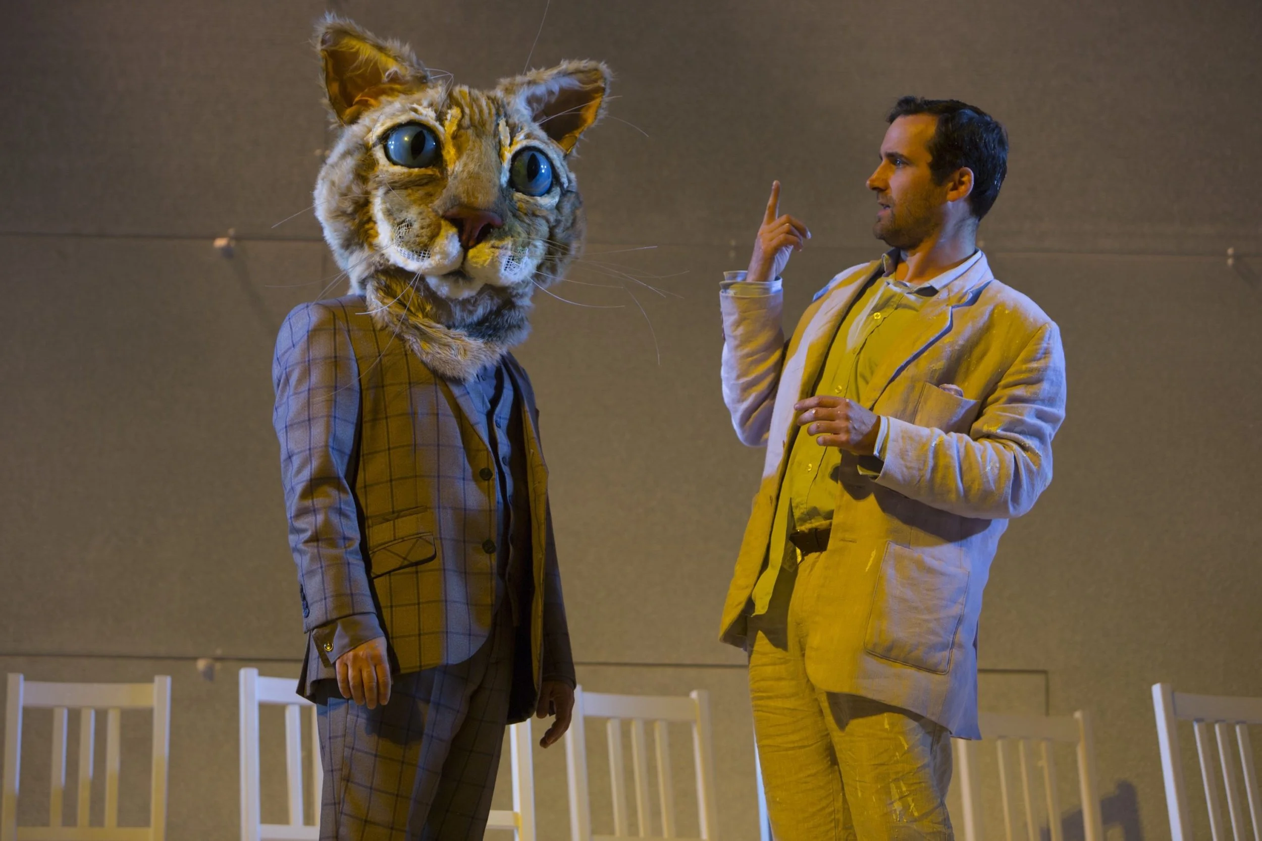 A person wearing a yellow suit and another person wearing a checkered suit with a large cat mask standing in a room with beige walls and white chairs.