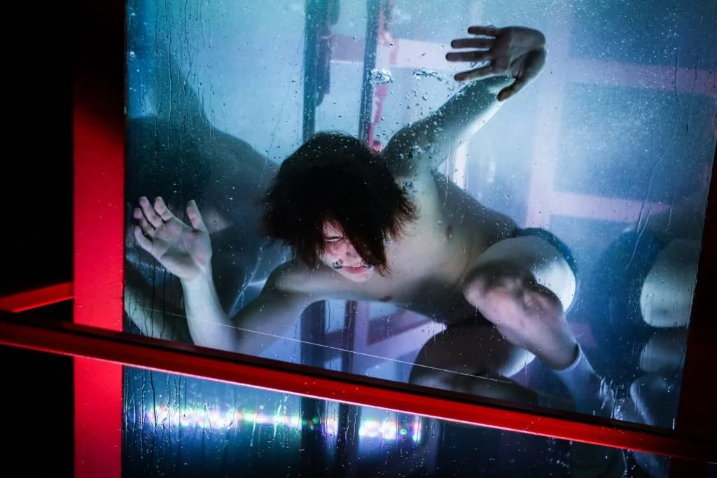 A woman with short dark hair and glasses lying on her side behind a glass panel with water droplets, her hand pressed against the glass and her eyes closed.