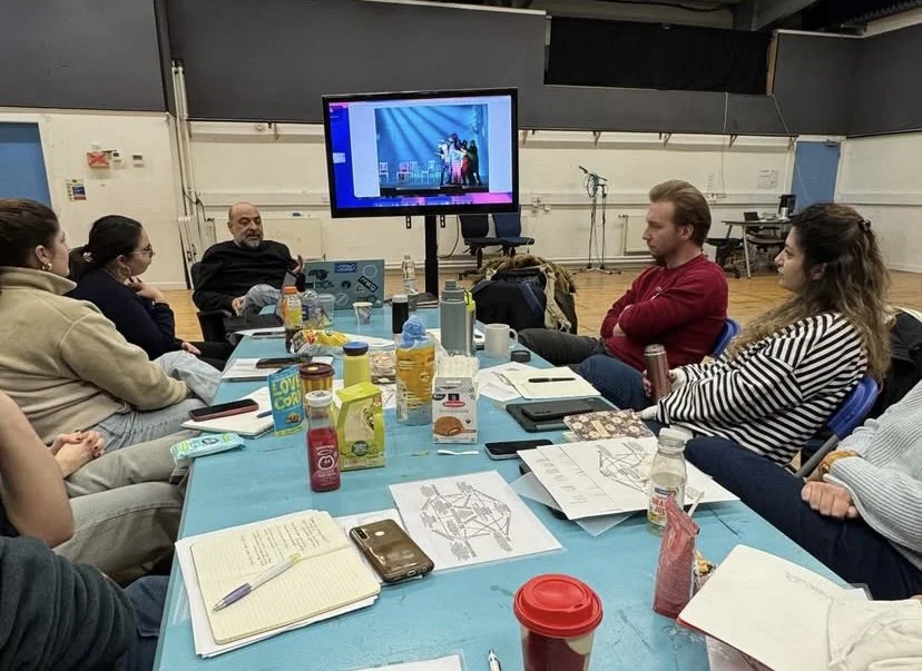 Group of people sitting around a conference table watching a presentation on a large screen, with various drinks, notebooks, and papers on the table.