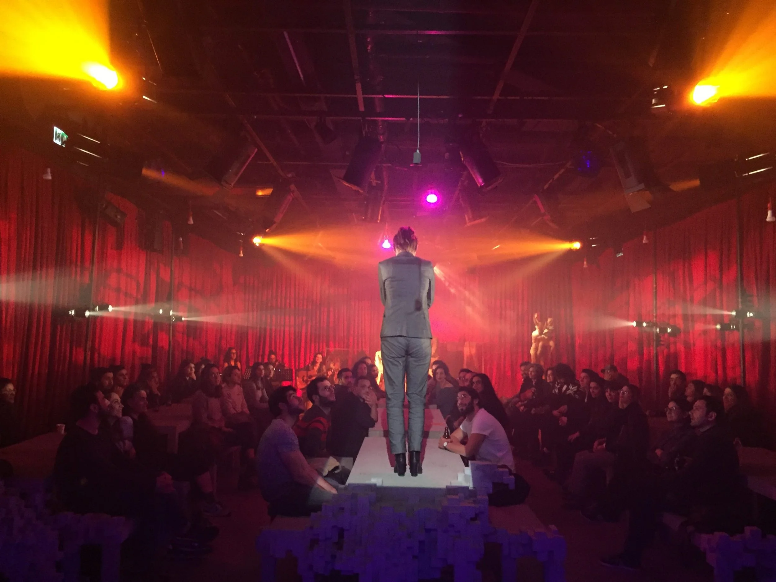 A person wearing a gray jacket and pants walking on a runway in front of an audience. The setting is a stage with red curtains and bright stage lights, with some yellow and purple lighting effects.