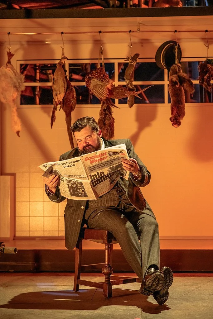 A man in a pinstripe suit with a beard is sitting on a wooden chair, reading a newspaper titled 'Socialist Worker.' The setting appears to be a stage with a backdrop of hanging hunted game animals and fur pelts, illuminated by warm, orange lighting.