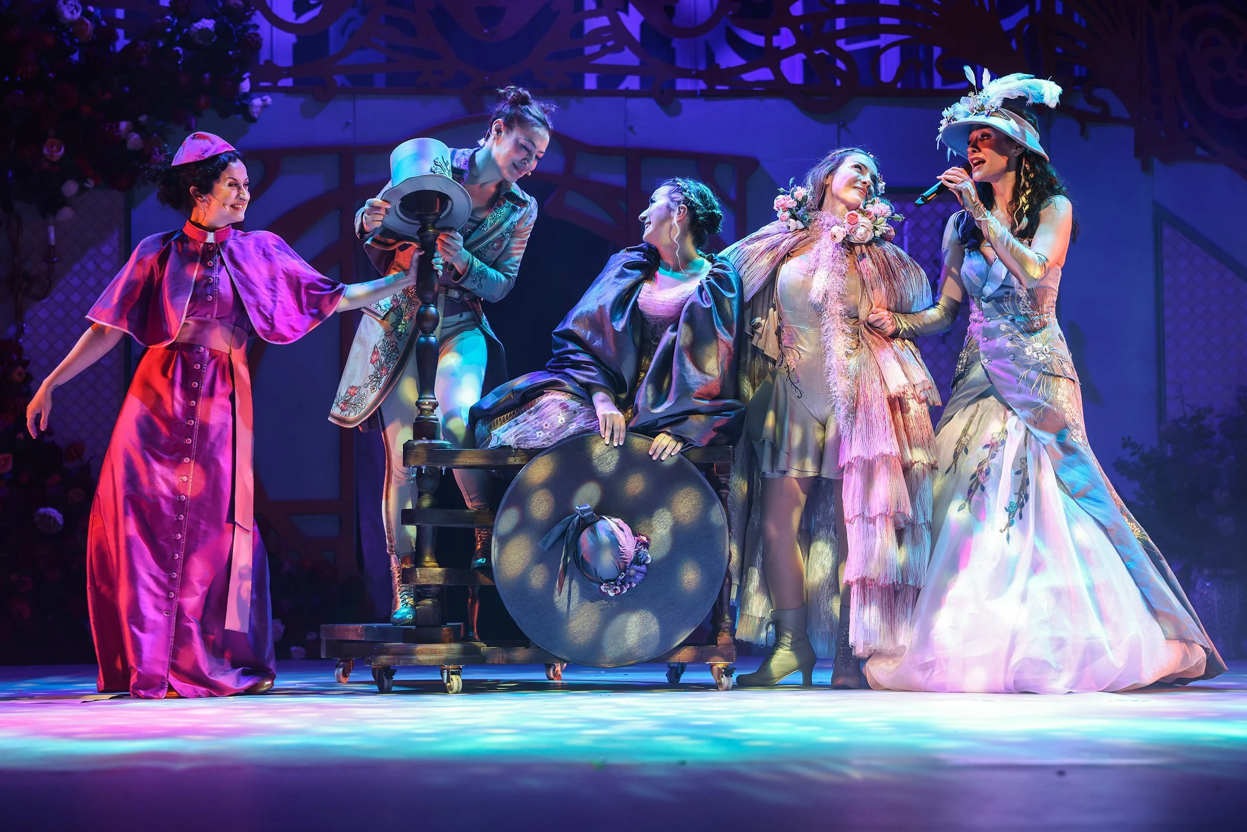 Actors performing on stage in colorful costumes, some seated and others standing, under vibrant lighting with a backdrop.