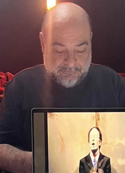 A man with a beard and bald head sitting at a table, looking down at a laptop. The laptop screen displays a person in a suit with an artistic, painted background.