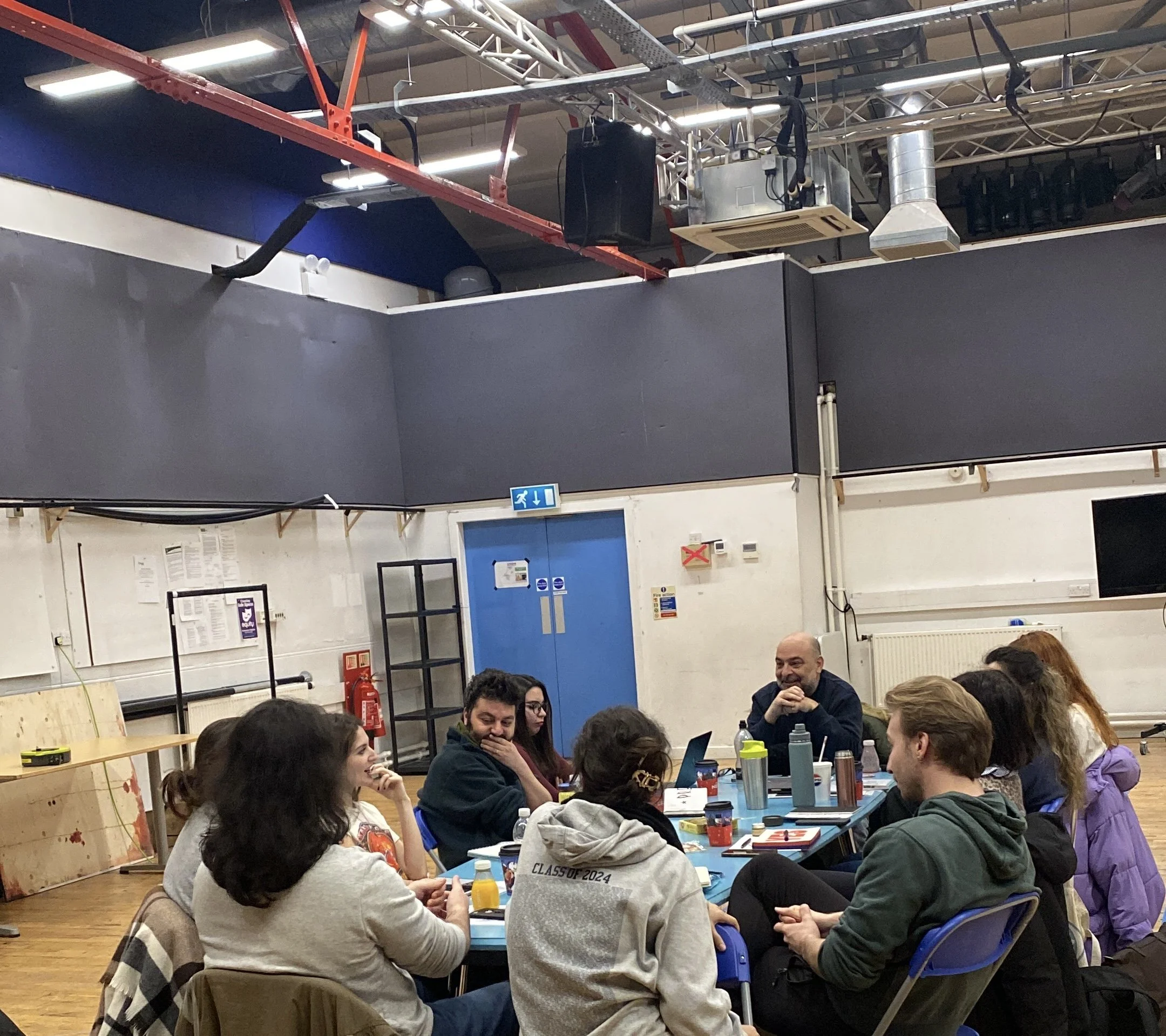 A group of people seated around a long table inside a community center or event hall, engaged in conversation and activities. The room has a high ceiling with exposed pipes and industrial lighting, with chairs, water bottles, and various snacks or it