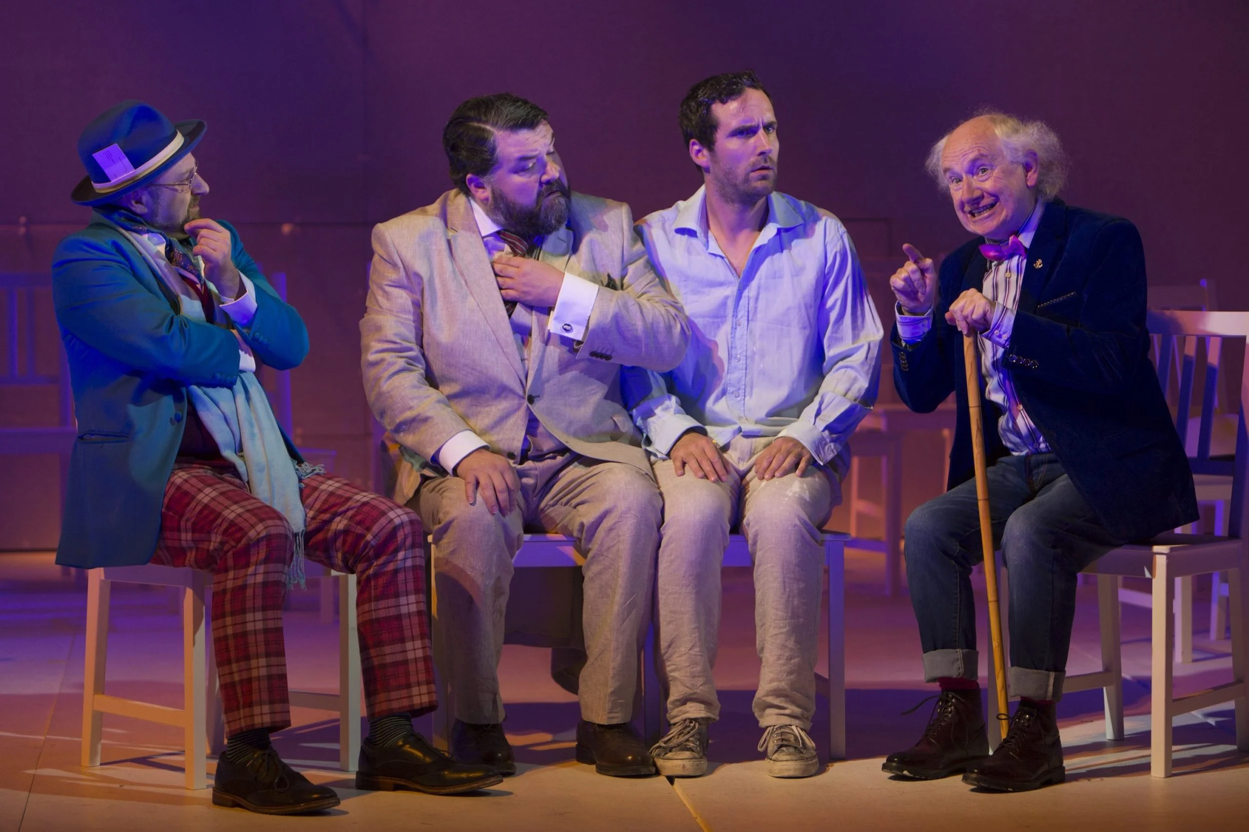 Five men sitting on chairs in a theatrical performance. The man on the far left wears a teal jacket, hat, and plaid pants, with his hand on his chin. The second man from the left has a beard and is dressed in a beige blazer and striped tie, holding h