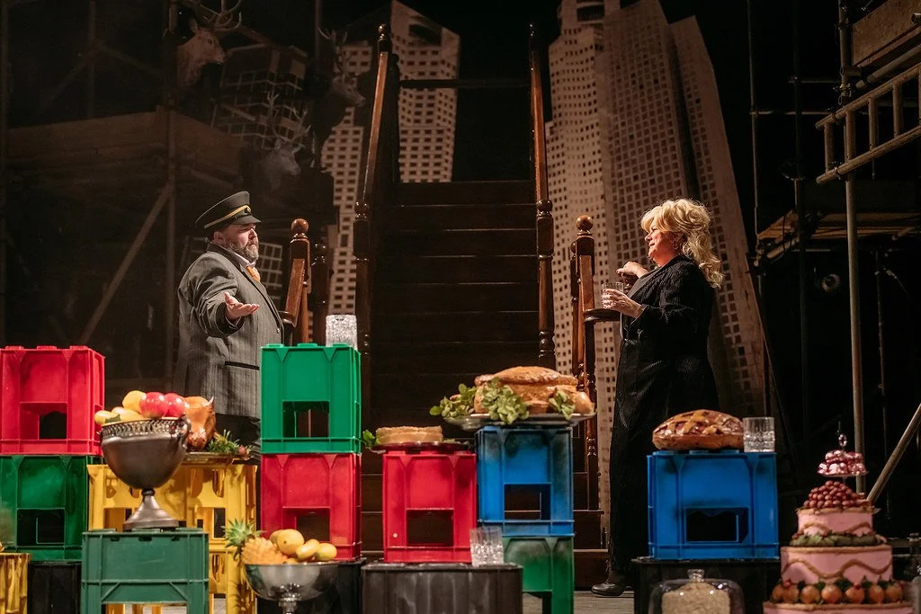 A theatrical stage scene with two actors, one man in a conductor's or train ticket seller's uniform and a woman in black, surrounded by colorful crates and food items like cakes, sandwiches, and fruit, with a cityscape backdrop and stage scaffolding.
