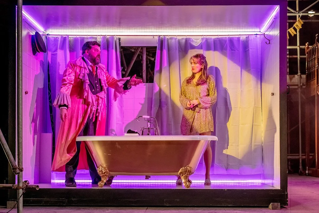 The image shows a theatrical scene with two actors on a stage set resembling a bathroom with a bathtub. The man on the left is dressed in an elaborate, colorful costume and appears to be speaking or performing, while the woman on the right, dressed i