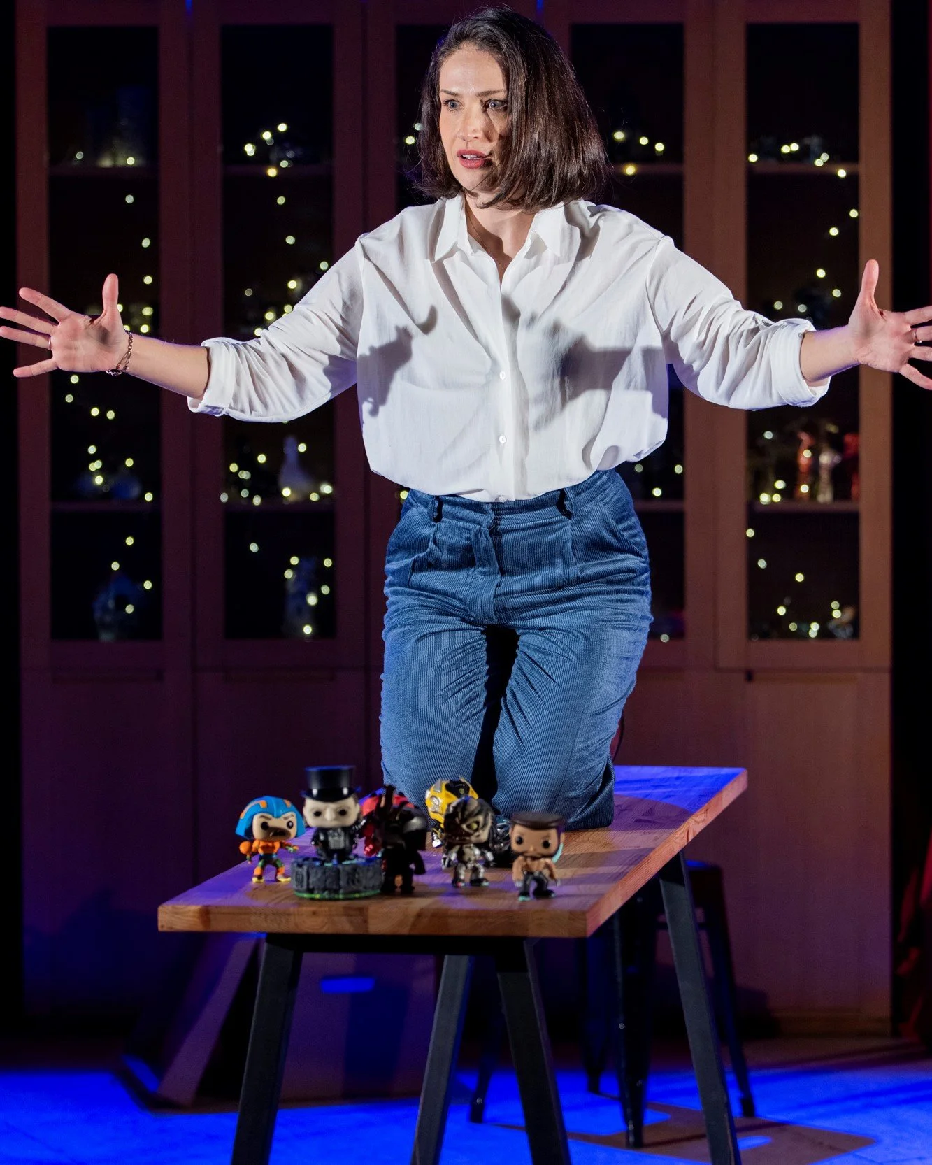 Woman in white shirt and blue jeans standing in front of a wooden table with various Funko Pop figurines, with shelves and string lights in the background.