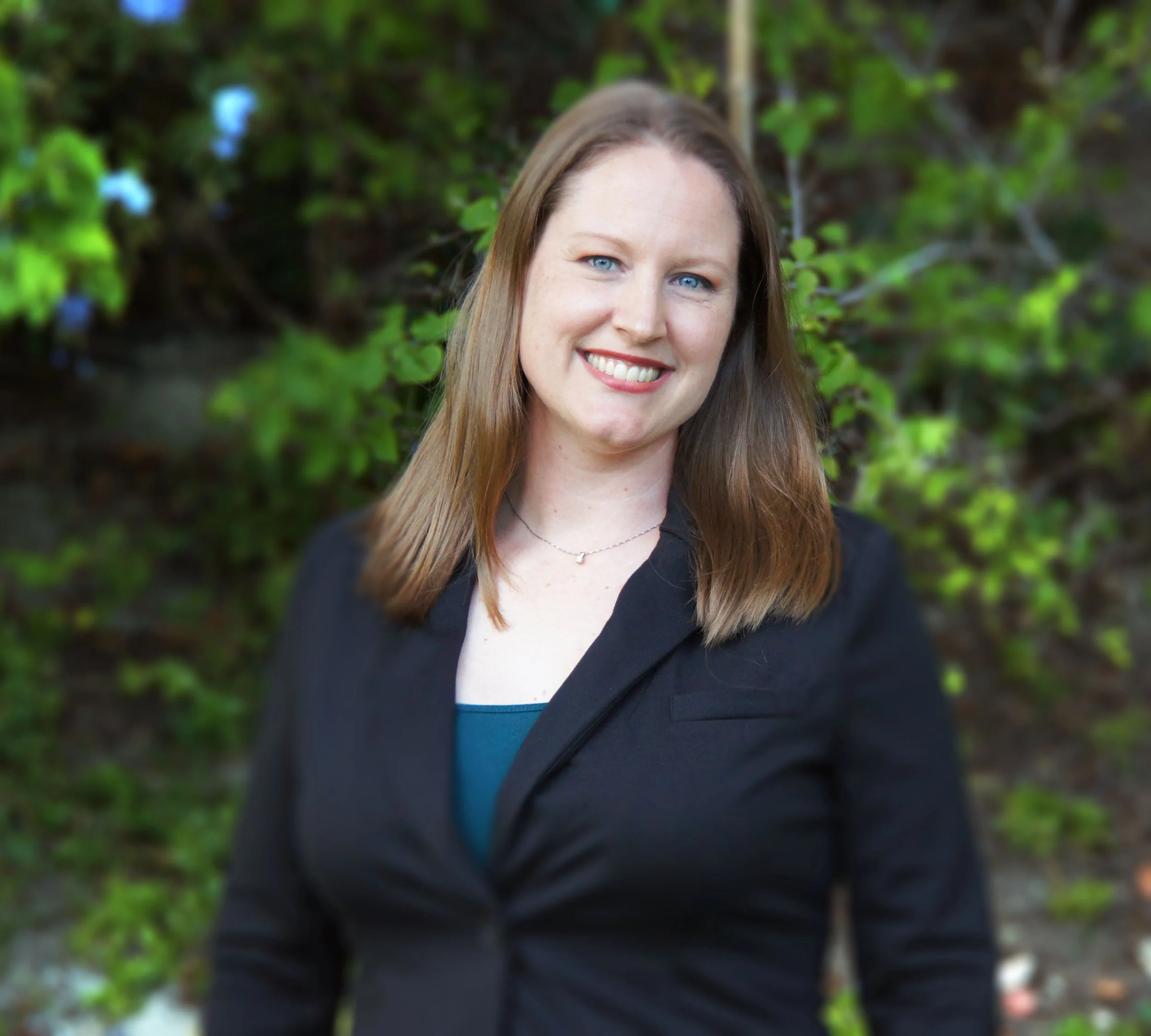 A woman with long brown hair and blue eyes smiling, wearing a black jacket and a necklace, standing outdoors with green foliage in the background.