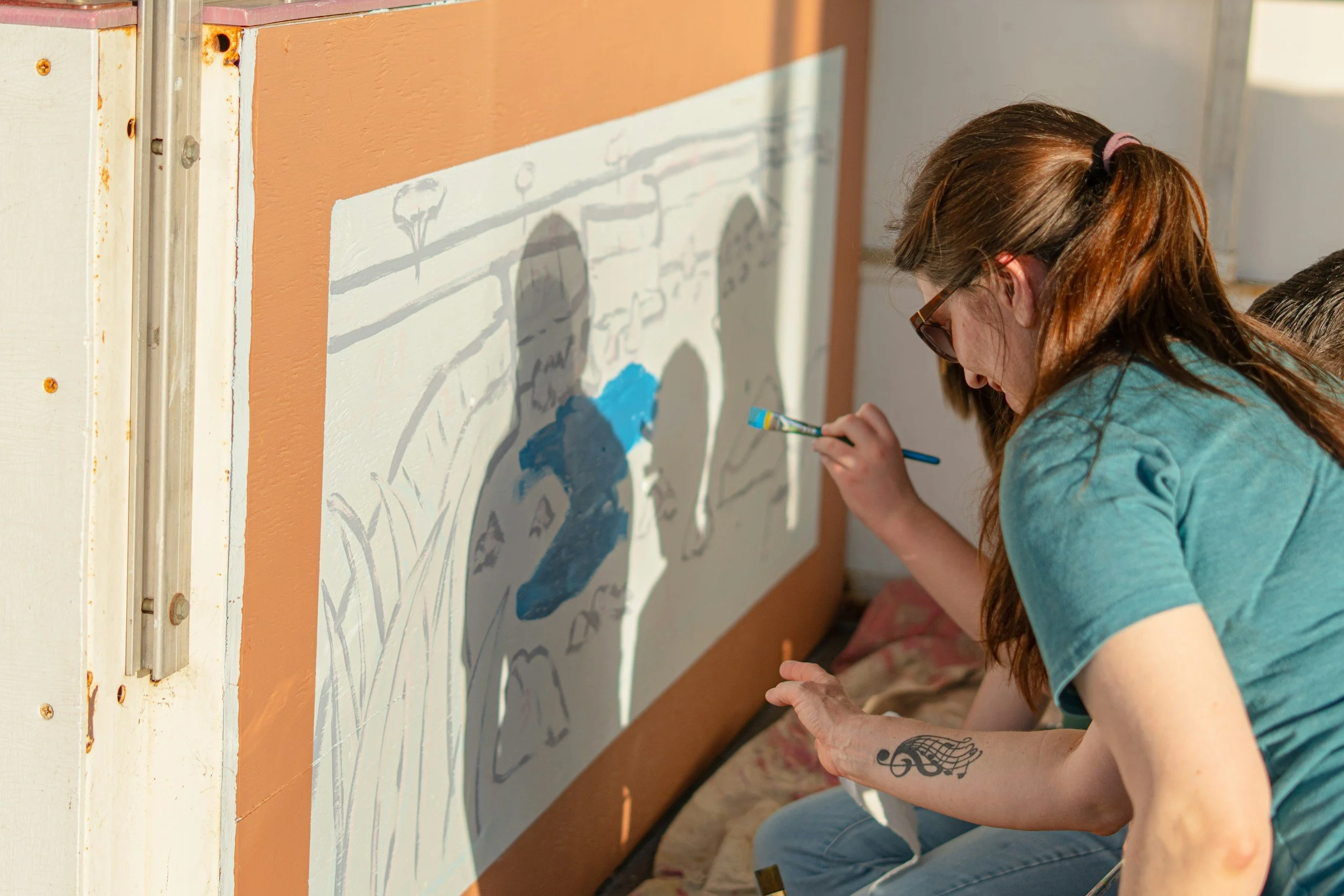 Person with long brown hair and sunglasses painting on a large canvas, with shadow of another person on the canvas, in a well-lit room.