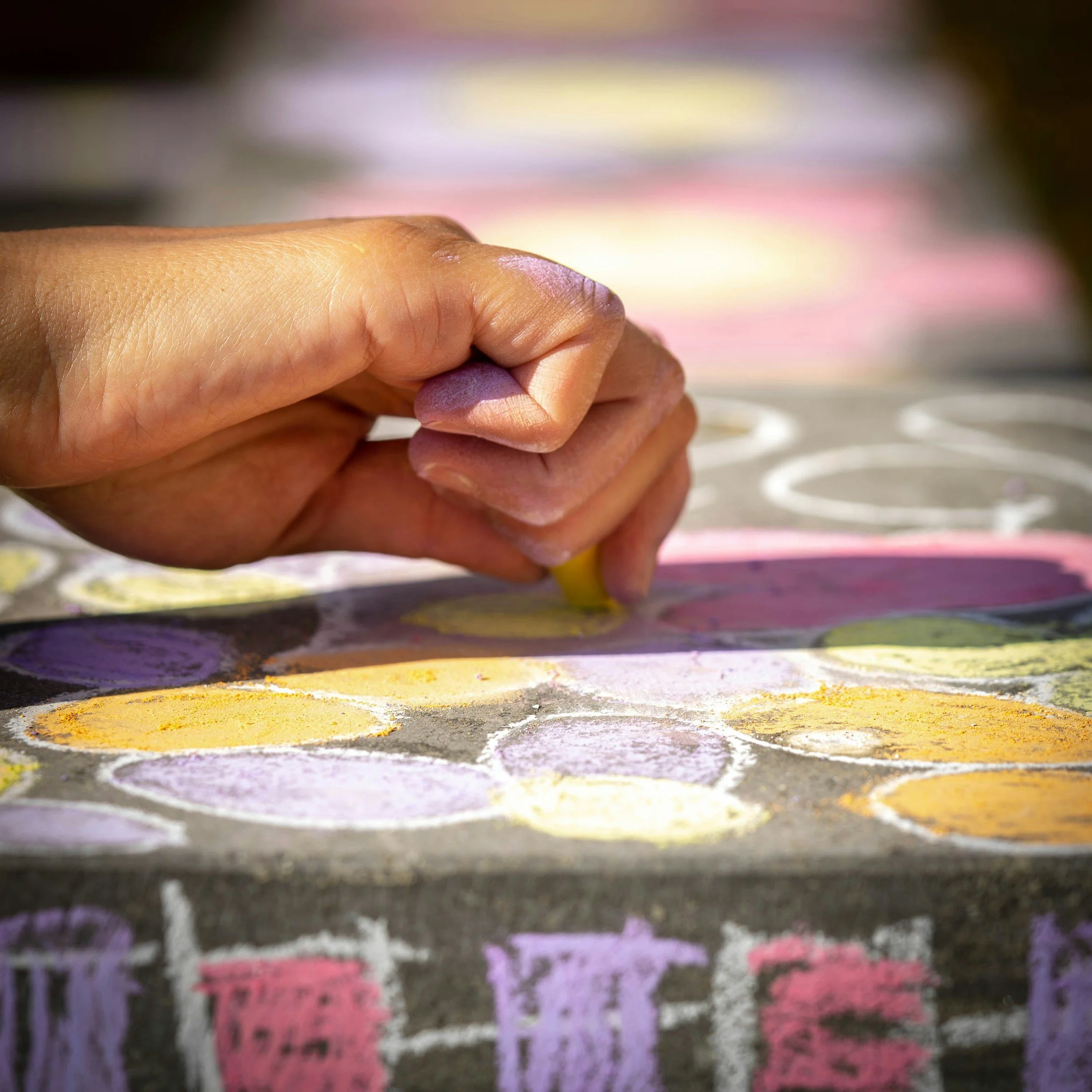 Close-up of a person's hand drawing on a colorful chalkboard with pastel chalks. The background is slightly blurred with vibrant chalk marks.
