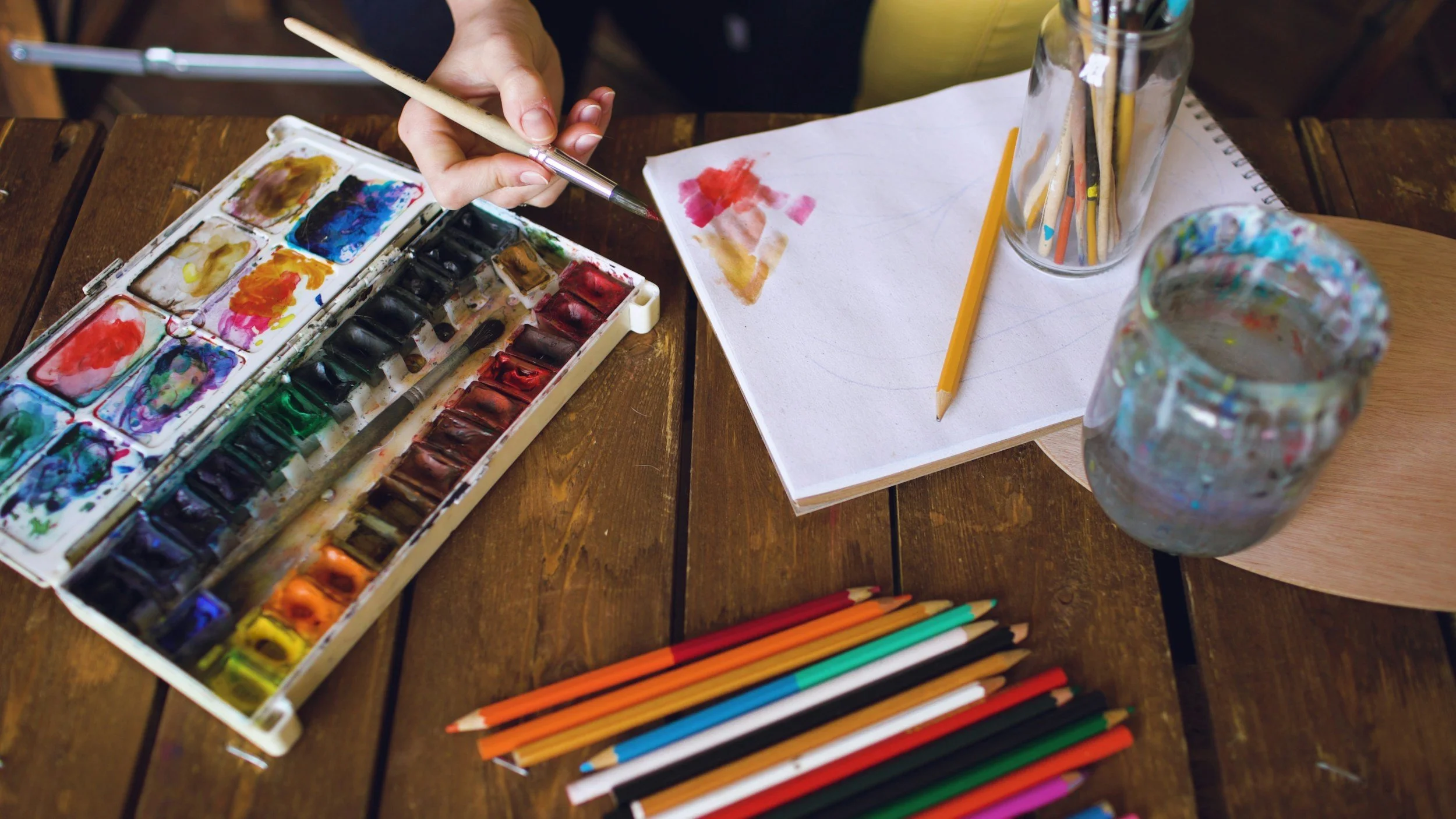 A person painting with watercolors on paper, with colored pencils, jars of brushes, and a watercolor palette on a wooden table.