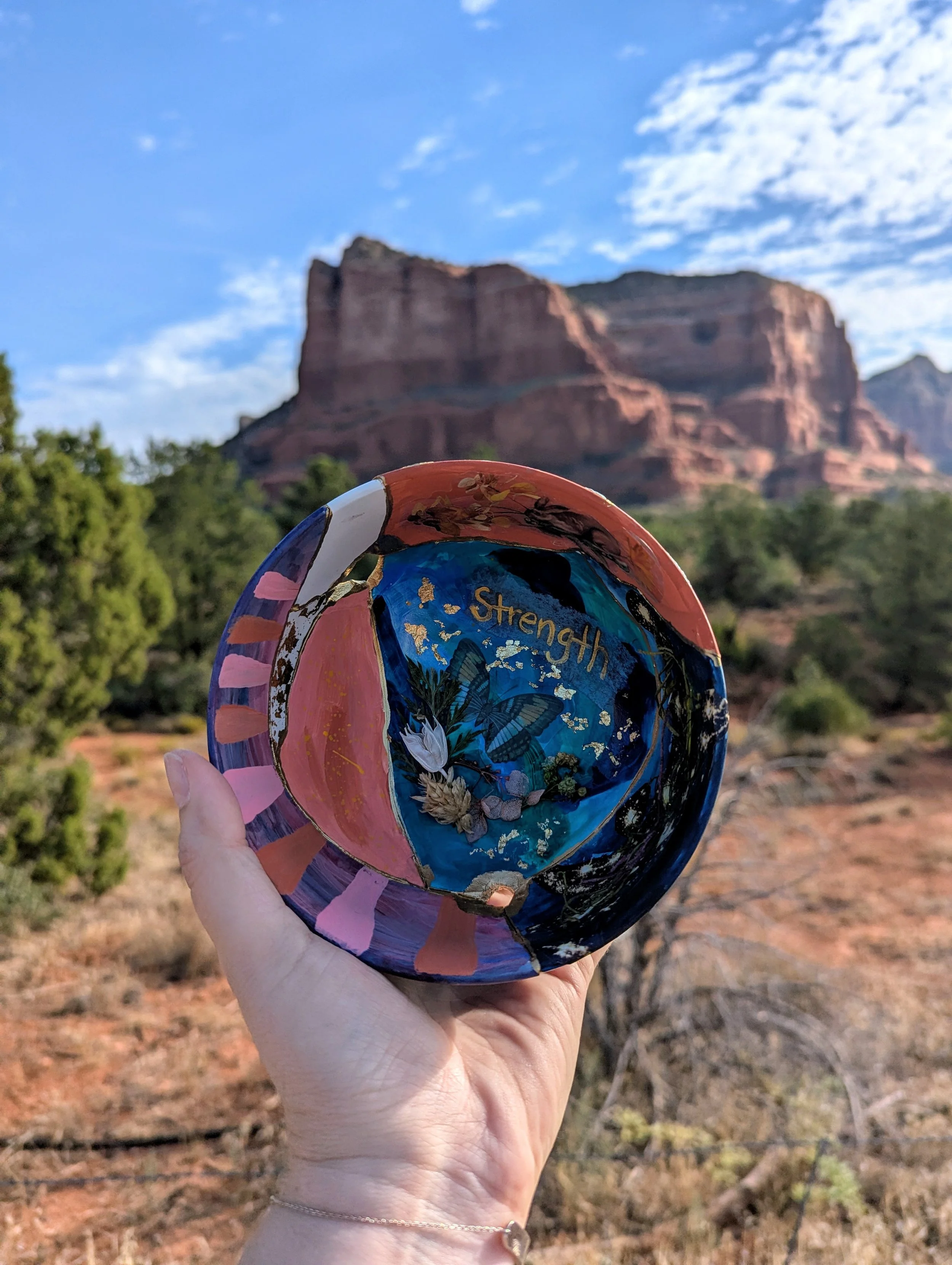 A hand holding a colorful painted bowl with a desert landscape and mountains in the background.
