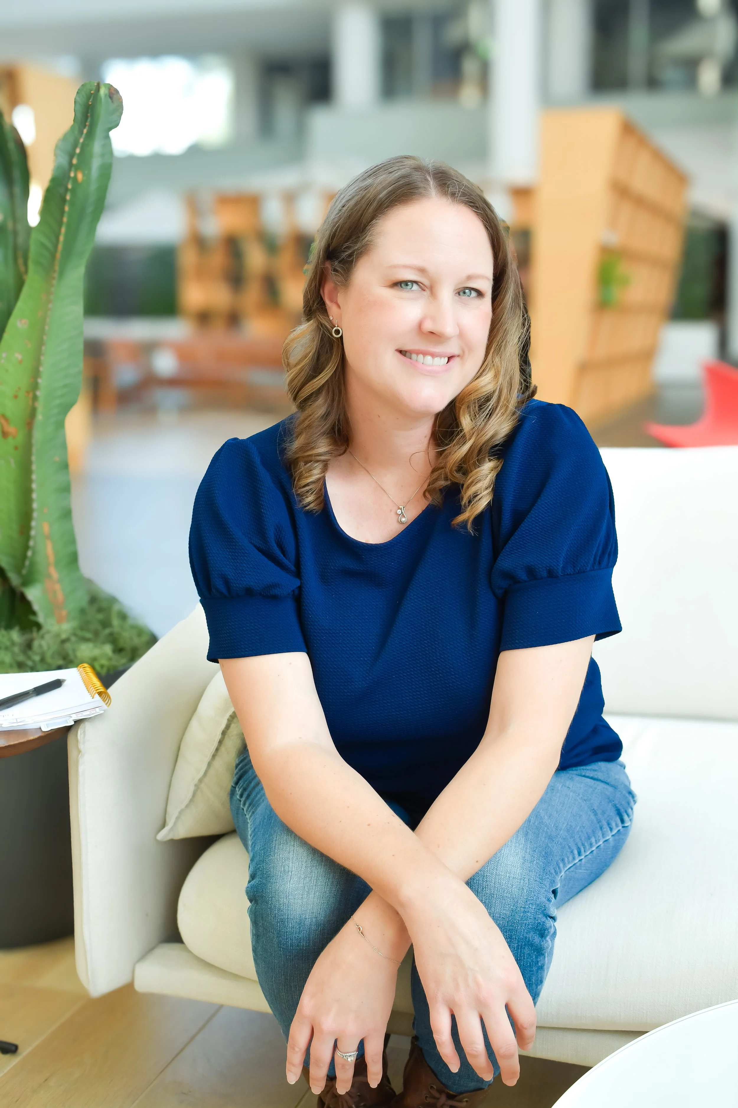 A woman with light skin, wavy brown hair, wearing a blue top, seated on a cream-colored armchair in a bright, modern indoor space. She is smiling and looking directly at the camera with her arms resting on her lap. There is a cactus plant on her left and a small table with a notebook and pen nearby.