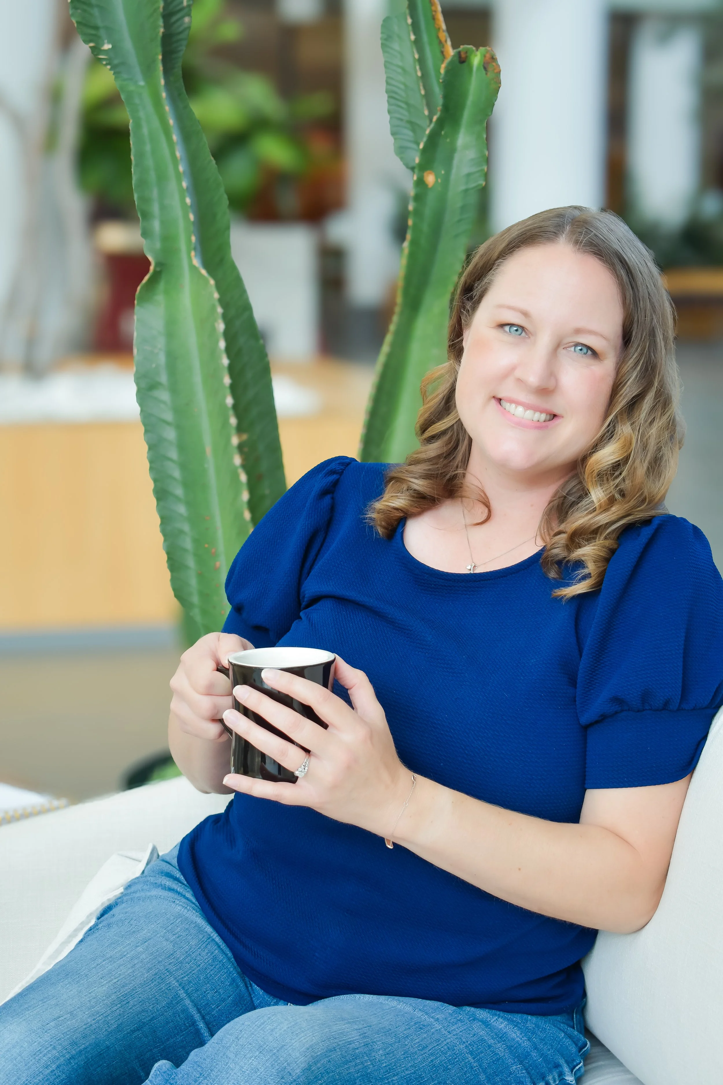A woman with wavy blonde hair and blue eyes smiling while holding a black and white mug. She is wearing a royal blue top with puffed sleeves and light blue jeans, sitting on a white couch next to a tall green cactus. The background appears to be an indoor space with plants and a blurred interior.