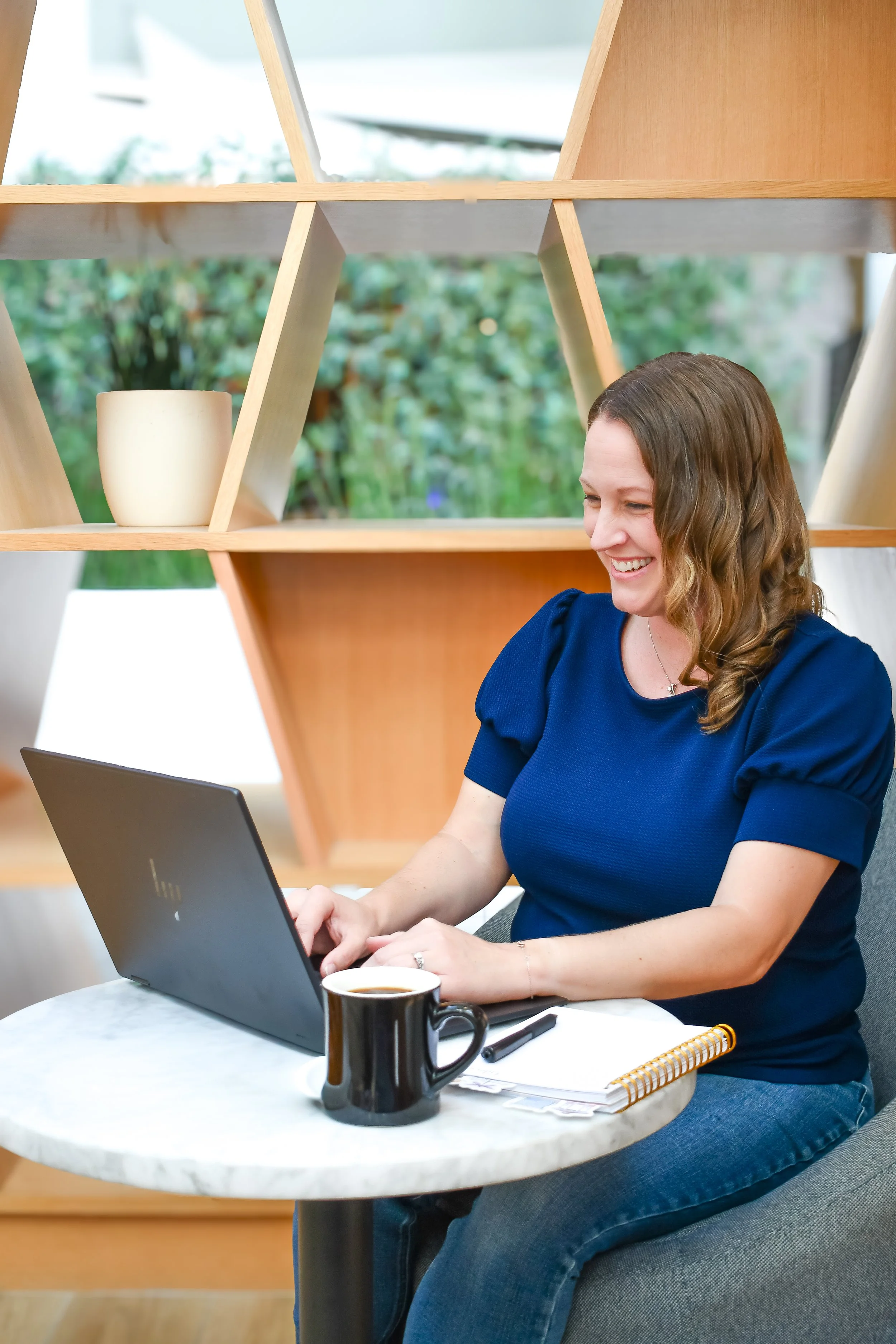 A woman sitting at a round table working on a laptop, with a coffee mug, notebook, and pen in front of her, and a background with a 3D wooden shelf and greenery outside a window.