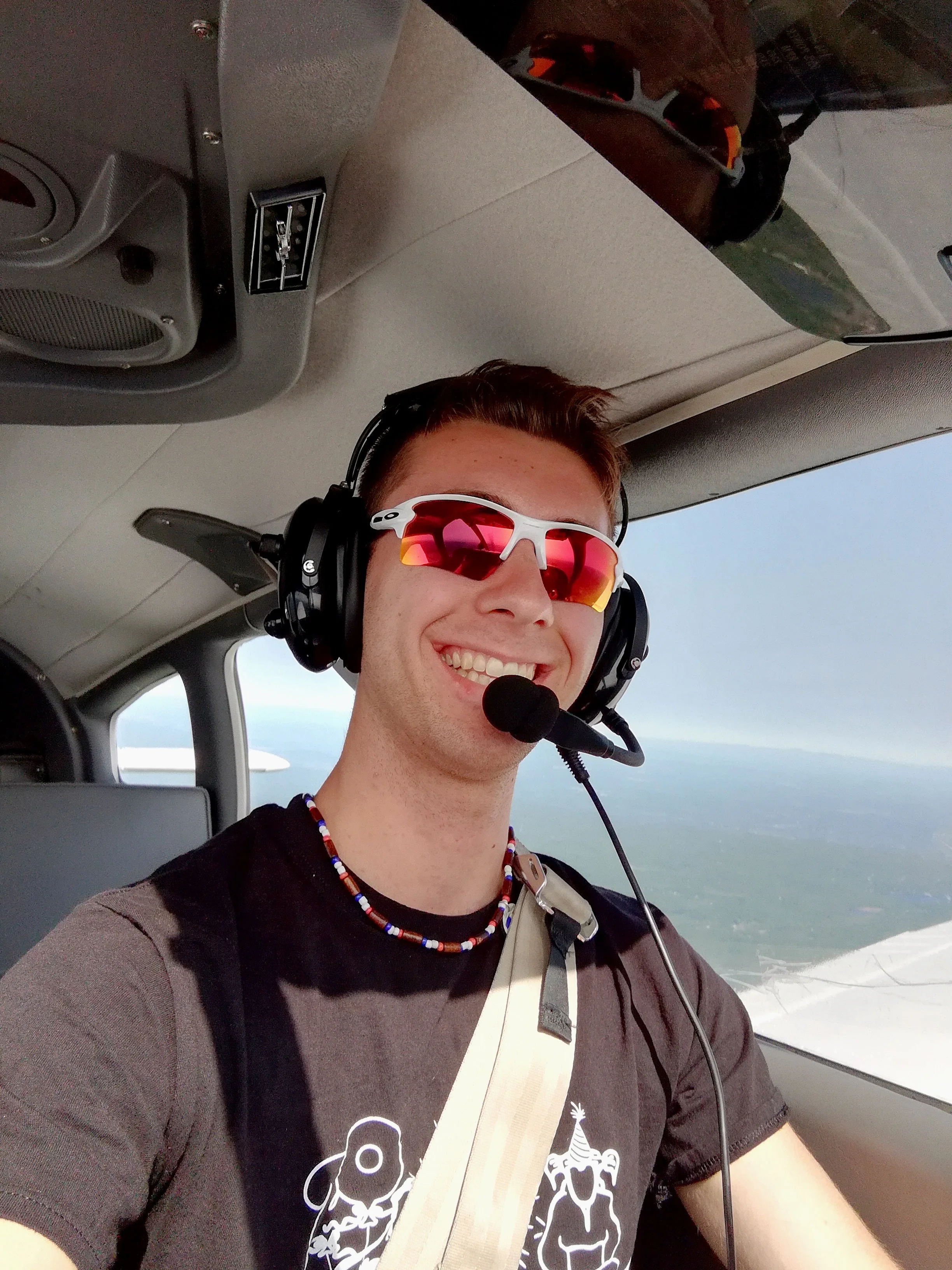 A smiling man wearing red-tinted sunglasses, a headset with a microphone, and a black T-shirt with white graphics, flying in a small aircraft over a landscape visible through the window.
