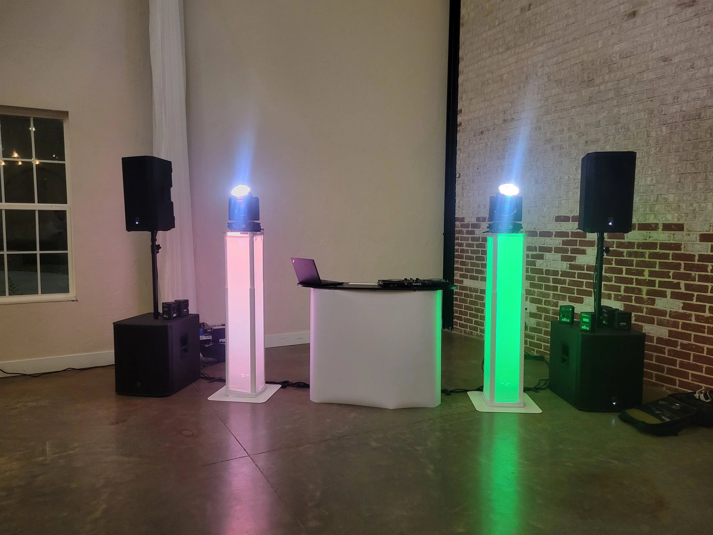 A DJ setup with speakers and colored LED light stands next to a brick wall in a decorated indoor space.