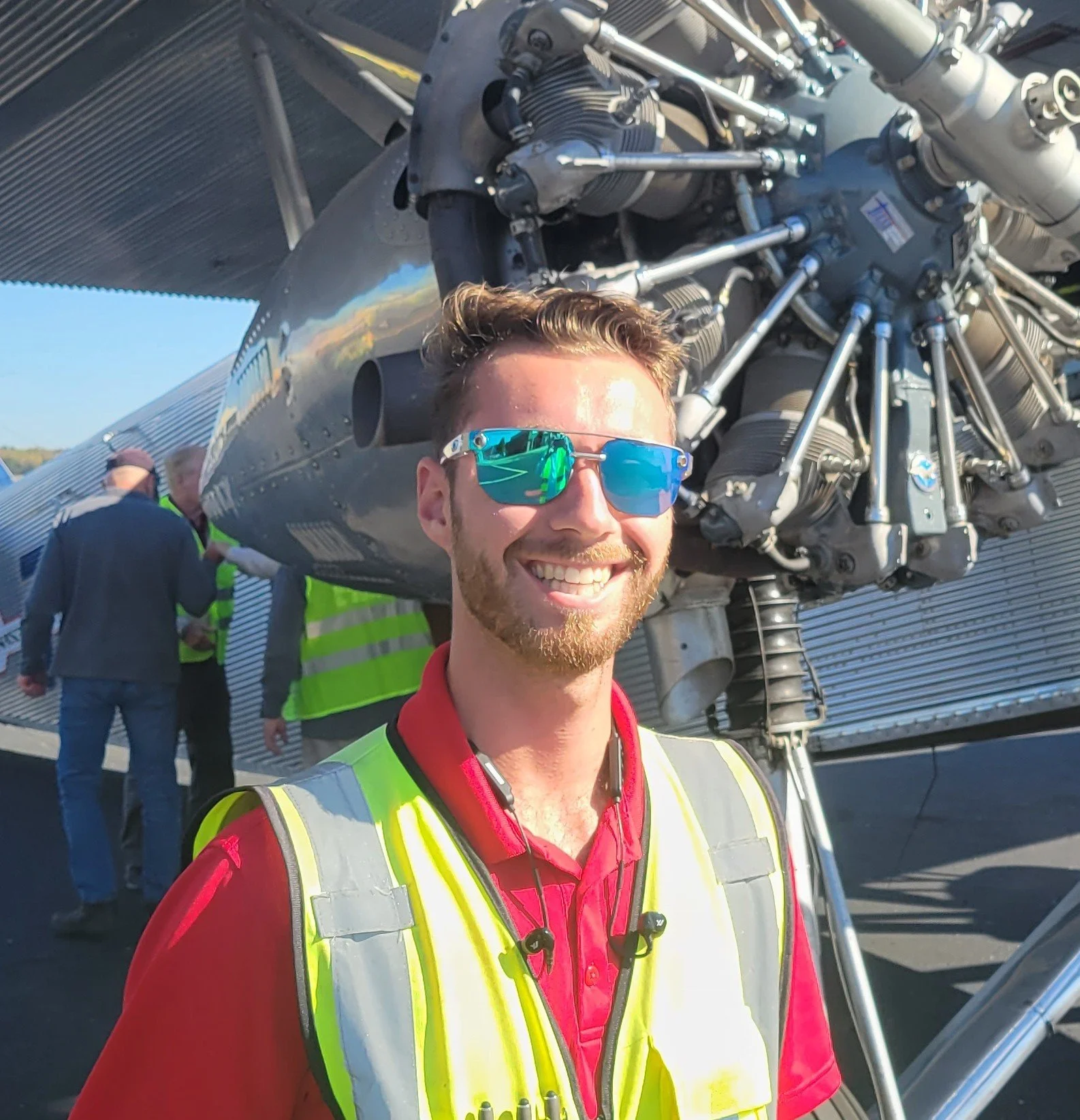 A young man with short hair and sunglasses smiling in front of a jet aircraft engine during an outdoor event. He's wearing a high-visibility safety vest over a red jacket.