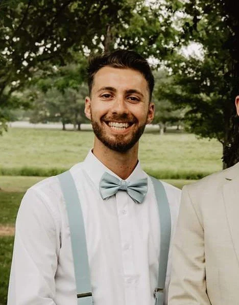 A smiling man with a beard, wearing a white shirt, light blue bow tie, and suspenders outdoors with trees and grass in the background.