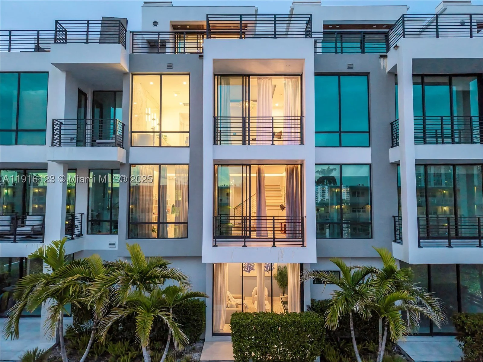 Modern multi-story apartment building with large glass windows and balconies surrounded by palm trees, illuminated from within.