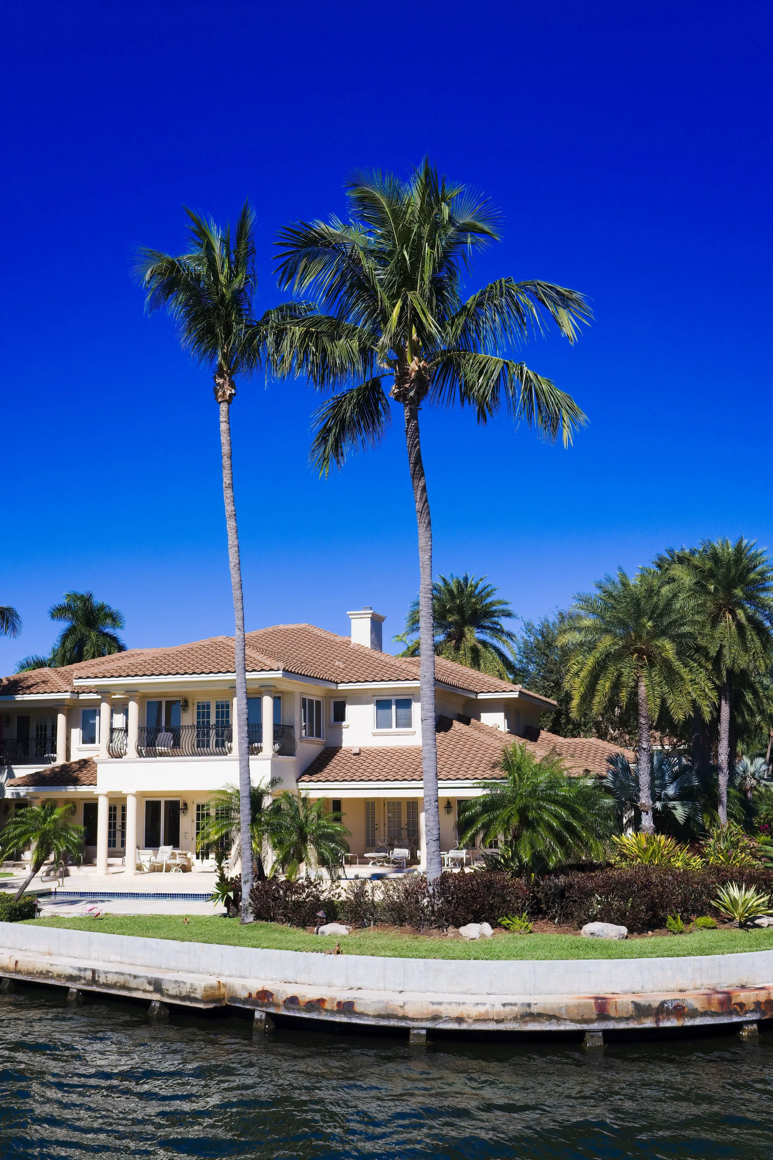 A large house with beige walls, red tile roof, and multiple balconies, surrounded by palm trees and greenery, located along a waterfront.