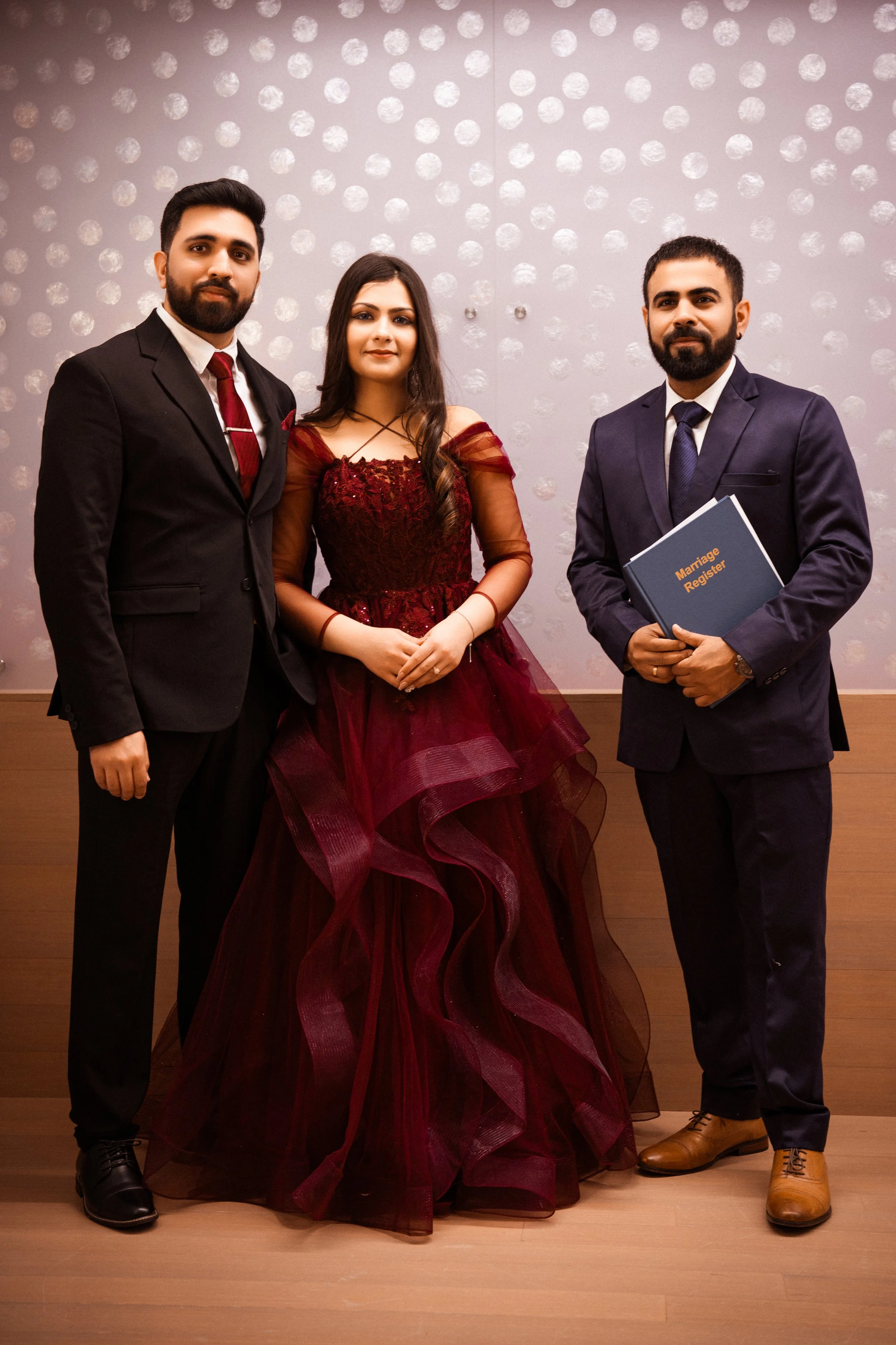 Three people in formal attire standing in front of a wall with decorative dots. The person on the right holds a blue folder labeled "Marriage Register."