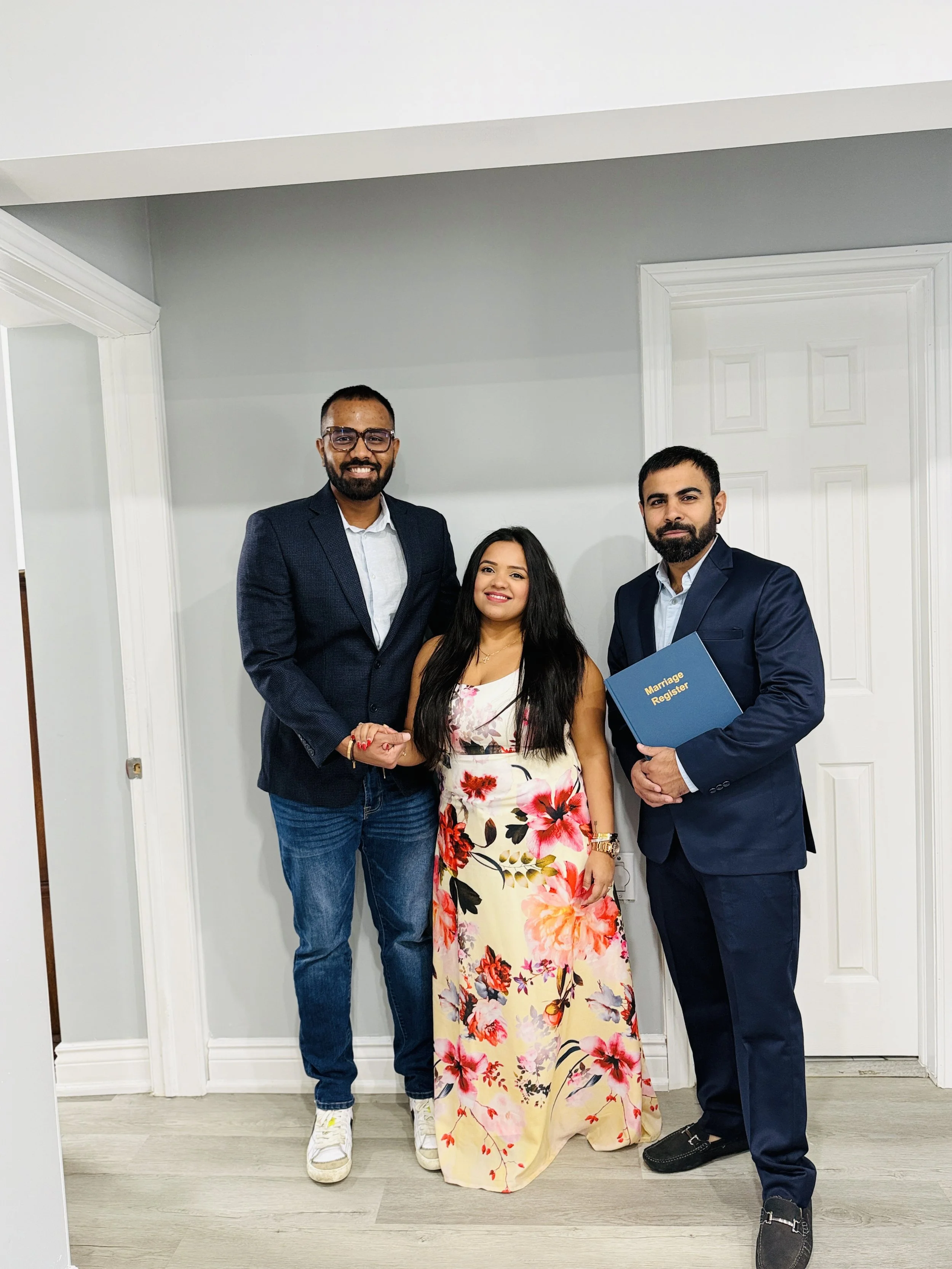 Three people standing in front of a white wall, two men and one woman. The woman is wearing a floral dress, and one man is holding a blue folder that says 'Marriage Register'.