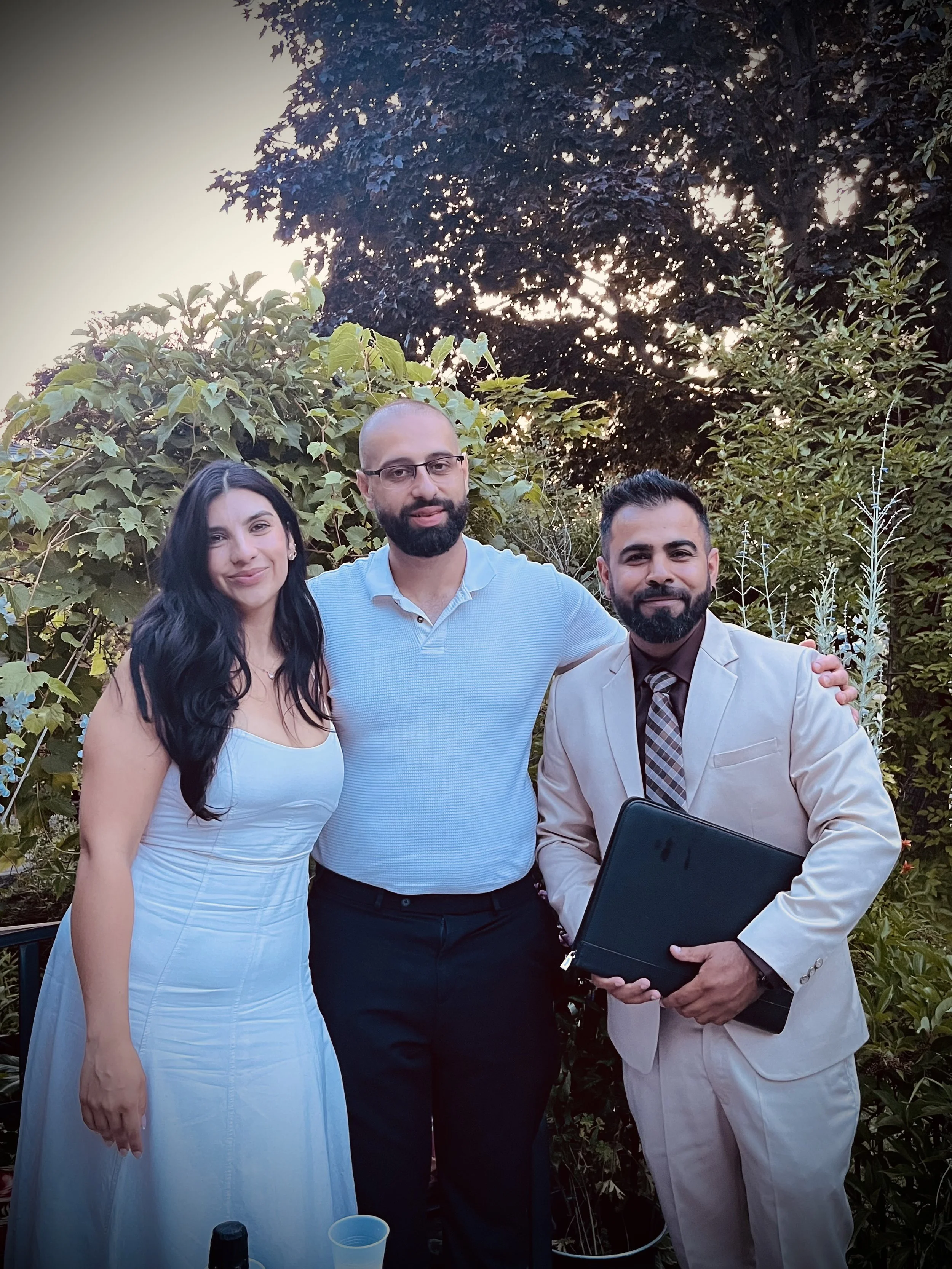 Three people posing outdoors at sunset, surrounded by greenery, with the woman on the left wearing a white dress, the man in the middle in a light blue polo shirt, and the man on the right in a beige suit holding a black folder.
