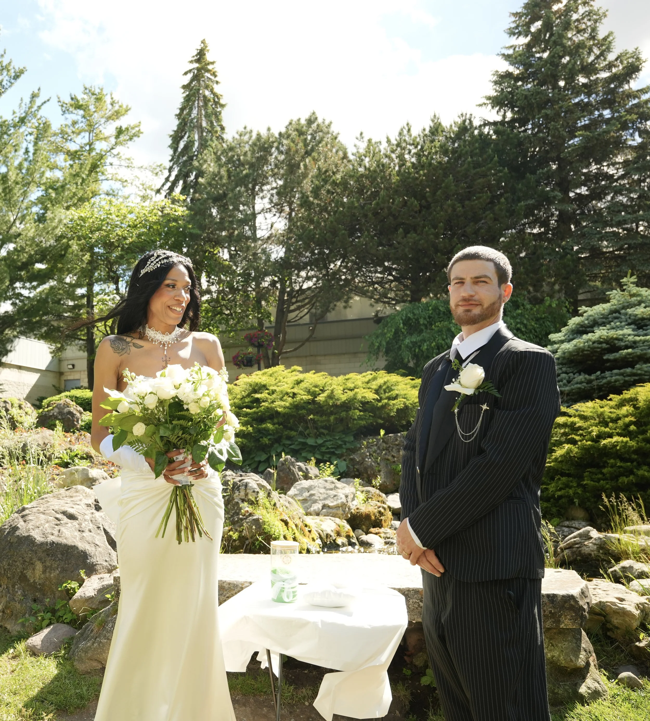 Couple standing at an outdoor wedding ceremony with their officiant's ceremony table, featuring a bride holding a white rose bouquet and a groom in a black pinstripe suit surrounded by lush gardens and natural stone landscaping.