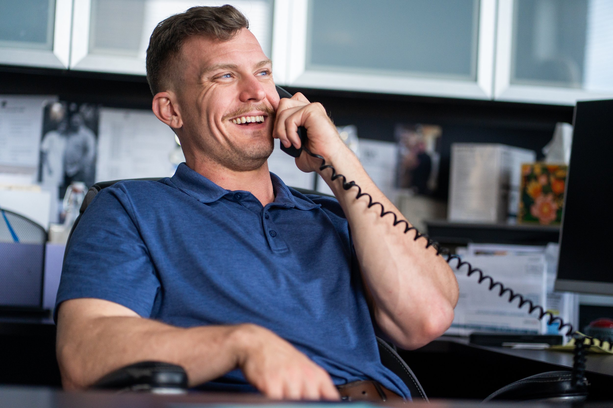 A man with short brown hair, blue eyes, and a beard is smiling while talking on a landline phone in an office setting. He is wearing a blue polo shirt and sitting at a desk with a computer, folders, and office supplies.