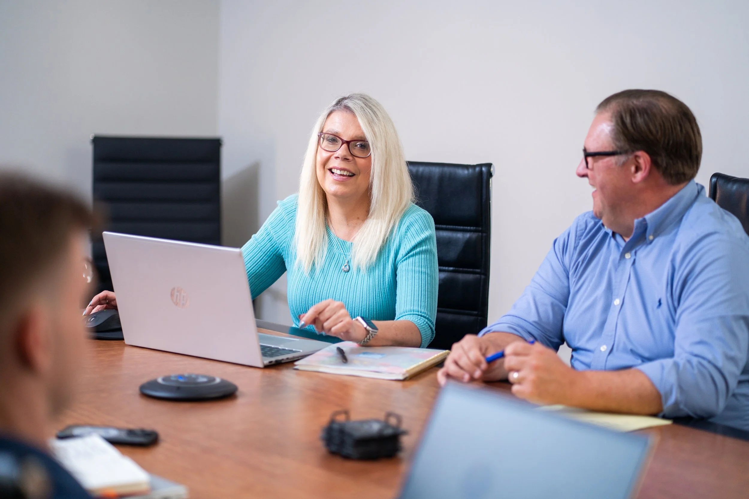 People sitting at a conference table in an office, engaged in a discussion, with a woman smiling and working on a laptop.