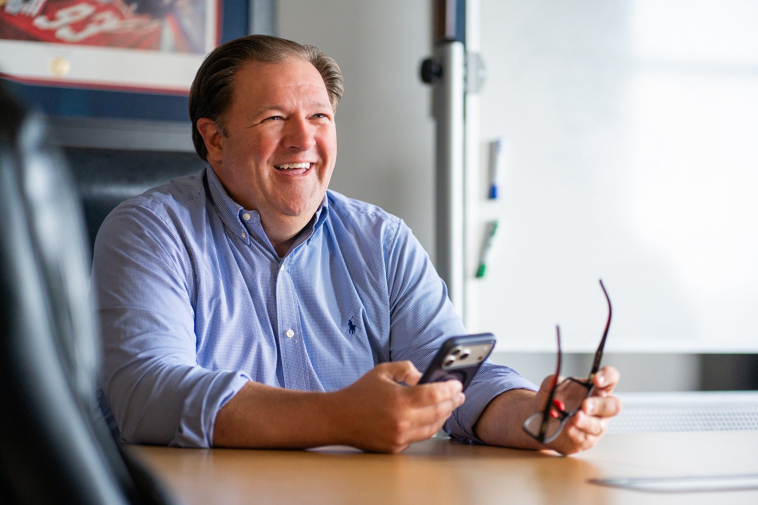 A smiling man with brown hair, wearing a light blue button-up shirt, sitting at a wooden table, looking at his phone.