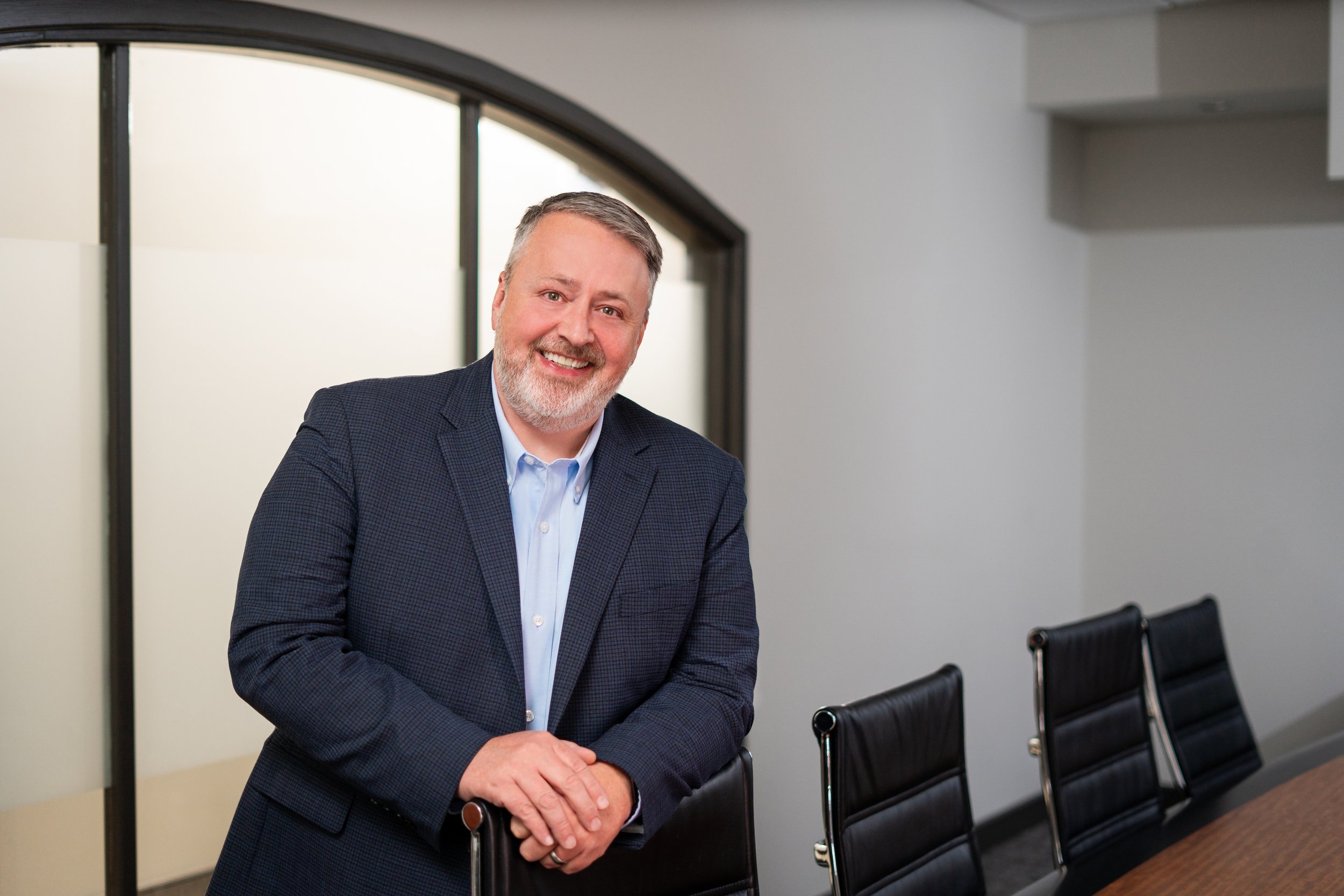 A smiling middle-aged man with gray hair and a beard, wearing a dark blue checkered blazer and light blue shirt, leaning on a conference table with black chairs in an empty meeting room.