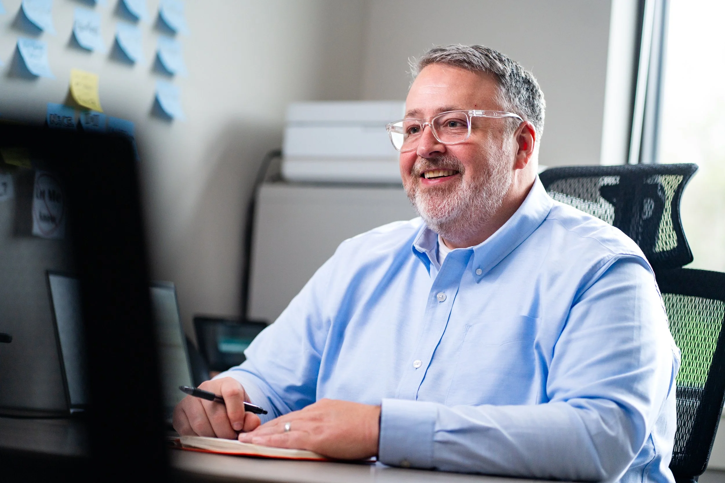 A smiling middle-aged man with gray hair and beard wearing clear glasses and a light blue shirt, sitting at a desk with a computer and holding a pen, in an office with a window and sticky notes on the wall.