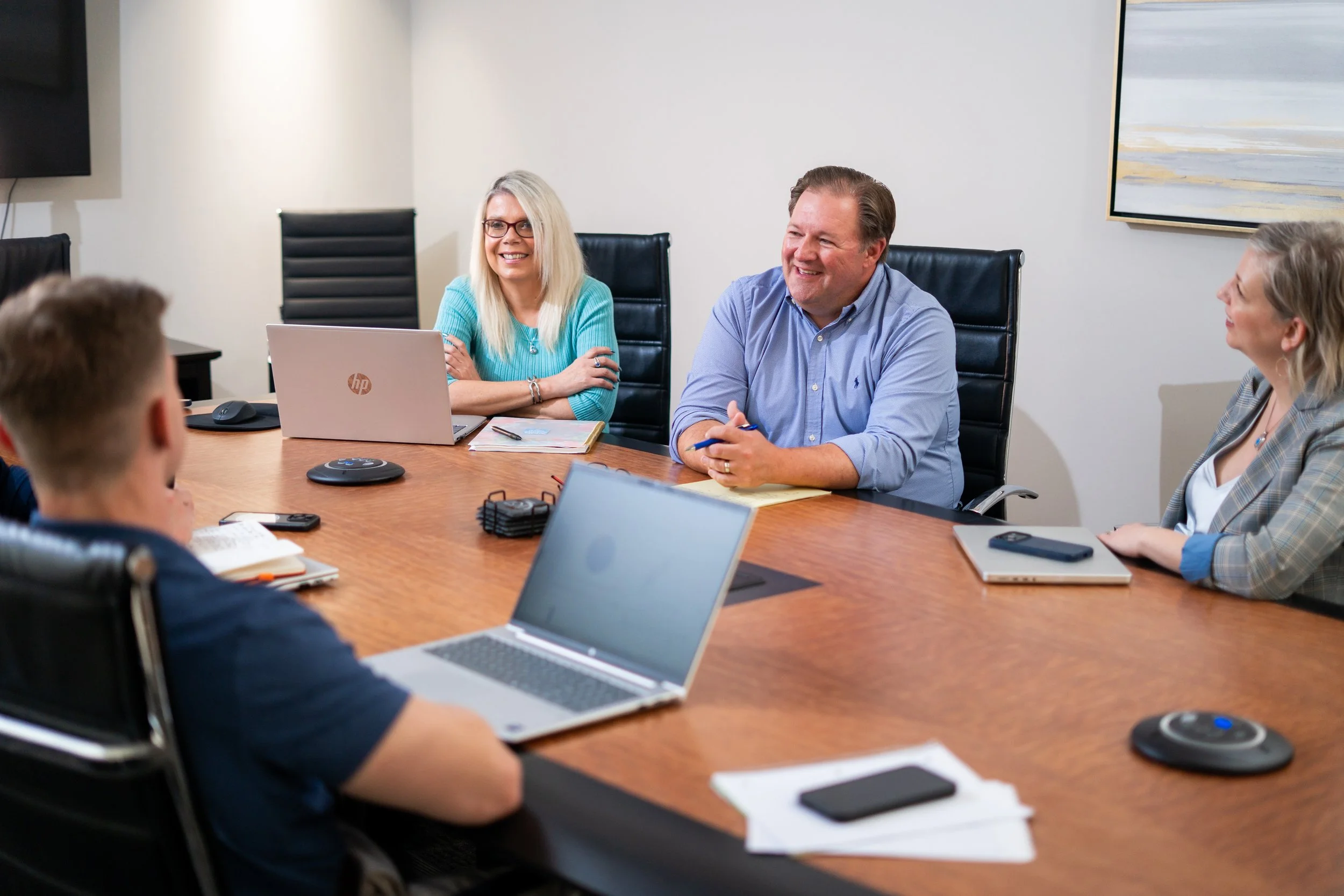 People sitting around a conference table engaged in a meeting, with laptops, notebooks, and conference call equipment on the table.