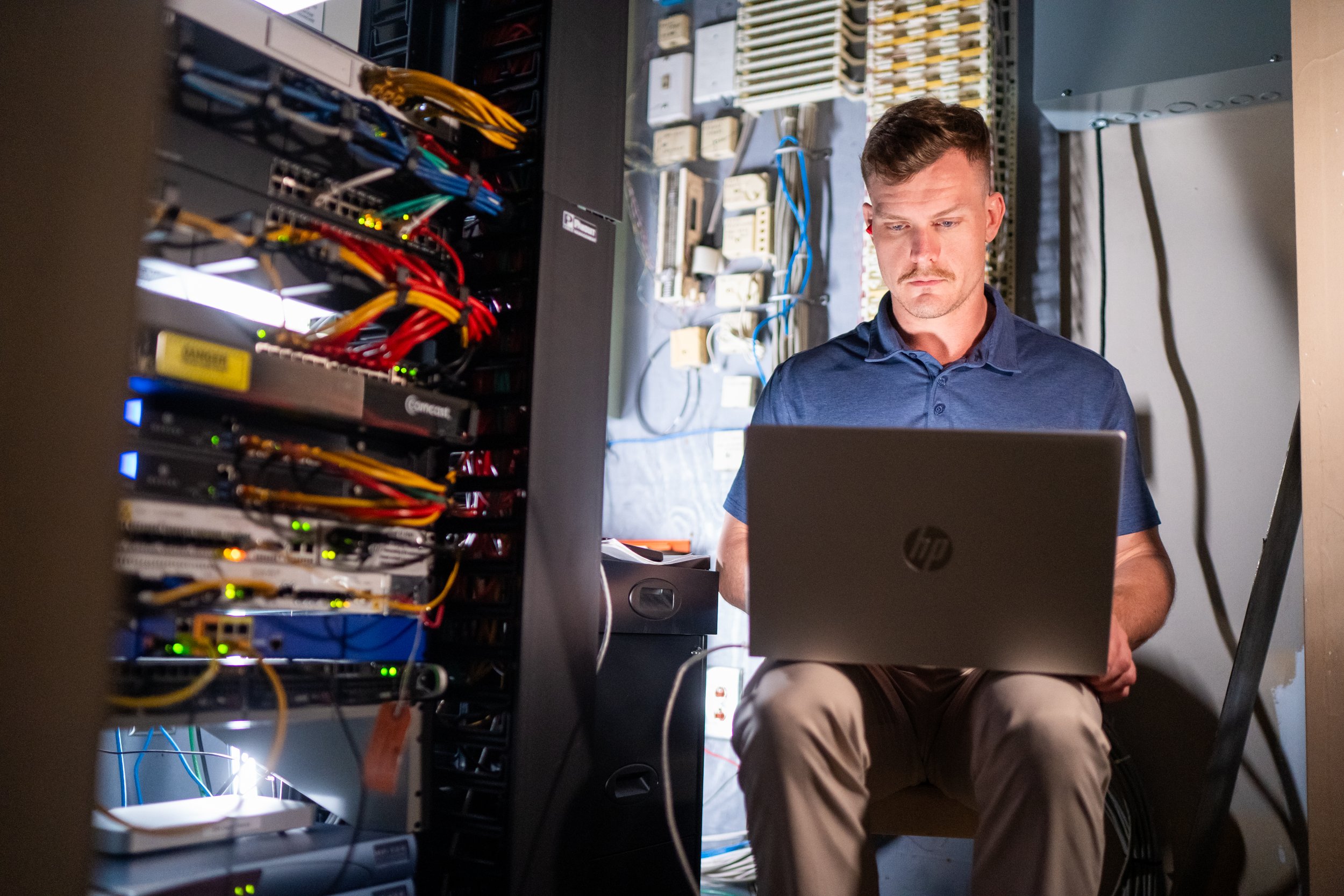 A man working on a laptop inside a server room with multiple rows of server racks and network cables.