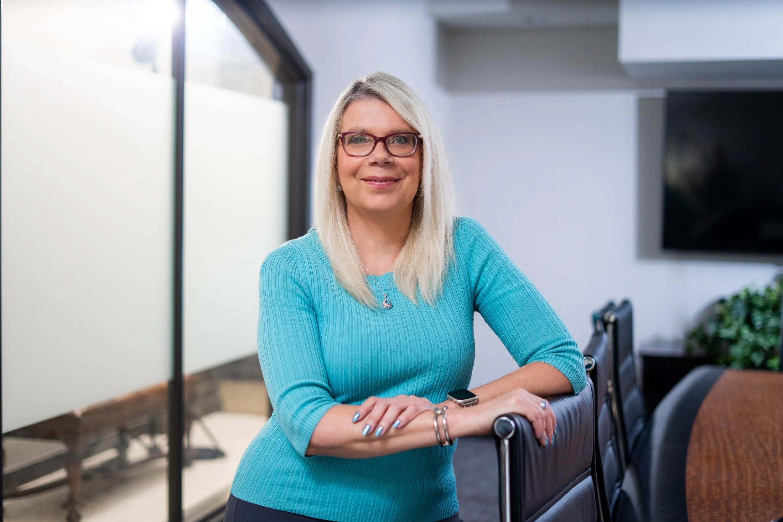 A middle-aged woman with blonde hair, glasses, and a turquoise sweater, smiling and sitting with her arms crossed in a modern office or conference room with white walls, large windows, and a tabletop.