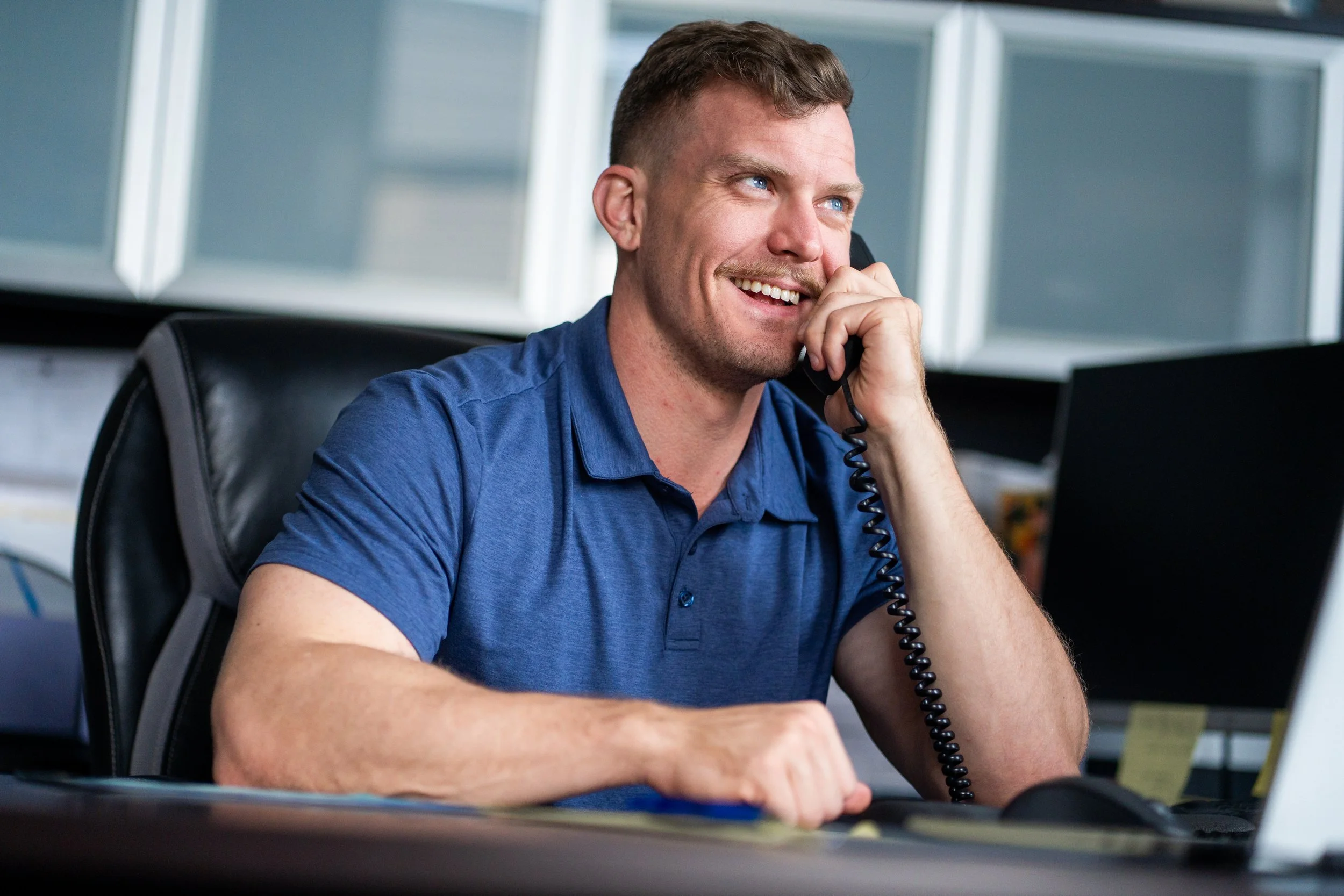 Smiling man in a blue shirt talking on a black corded phone in an office setting.