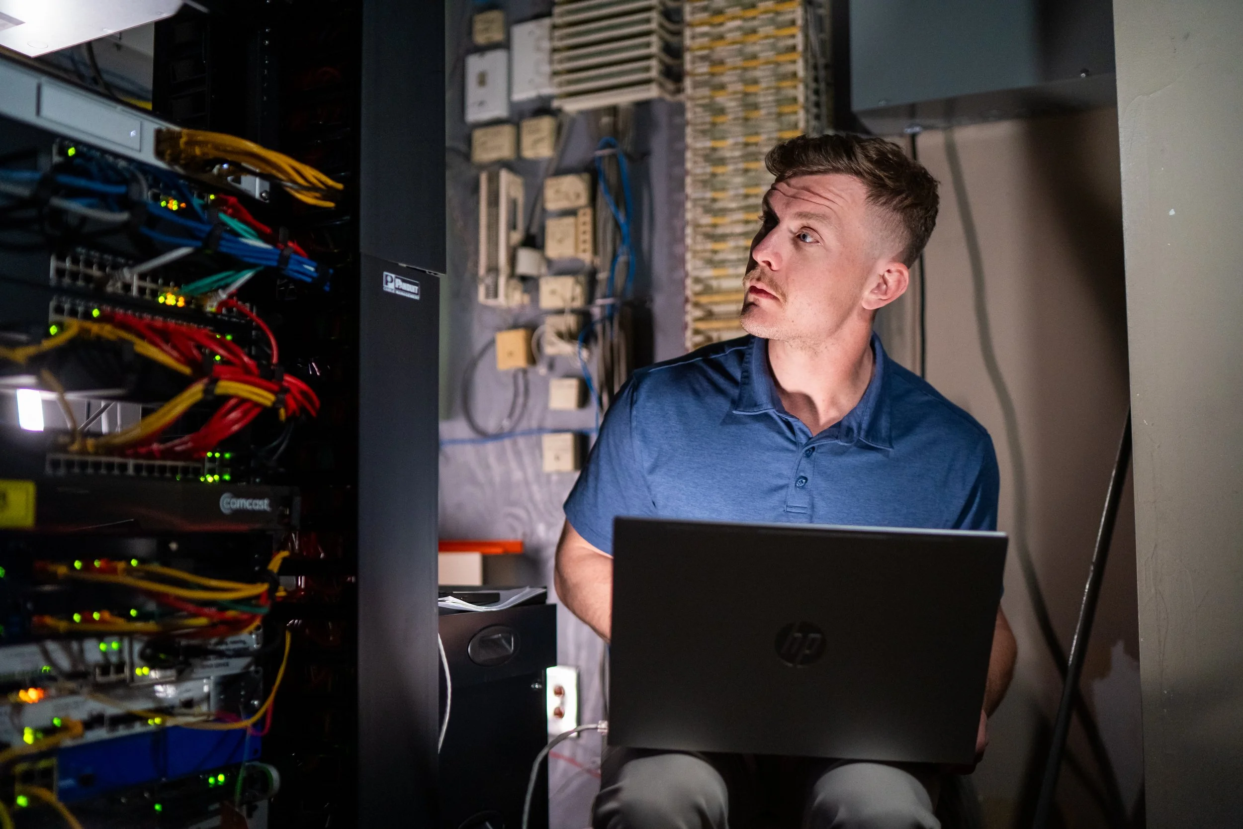 A man working in a data center or server room, seated in front of a server rack with various colored cables and blinking lights, focused on a laptop.