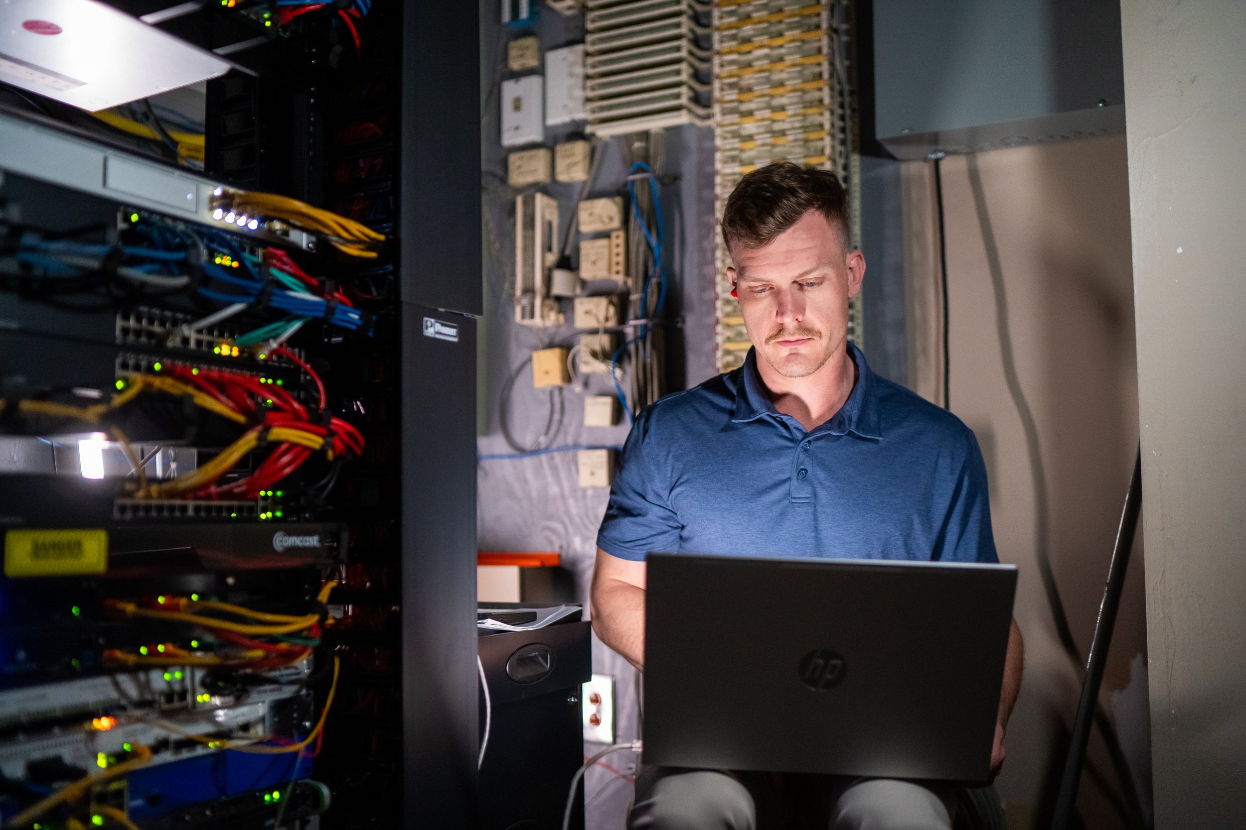 A man in a blue shirt working on a laptop in a server room, with server racks and networking equipment around him.