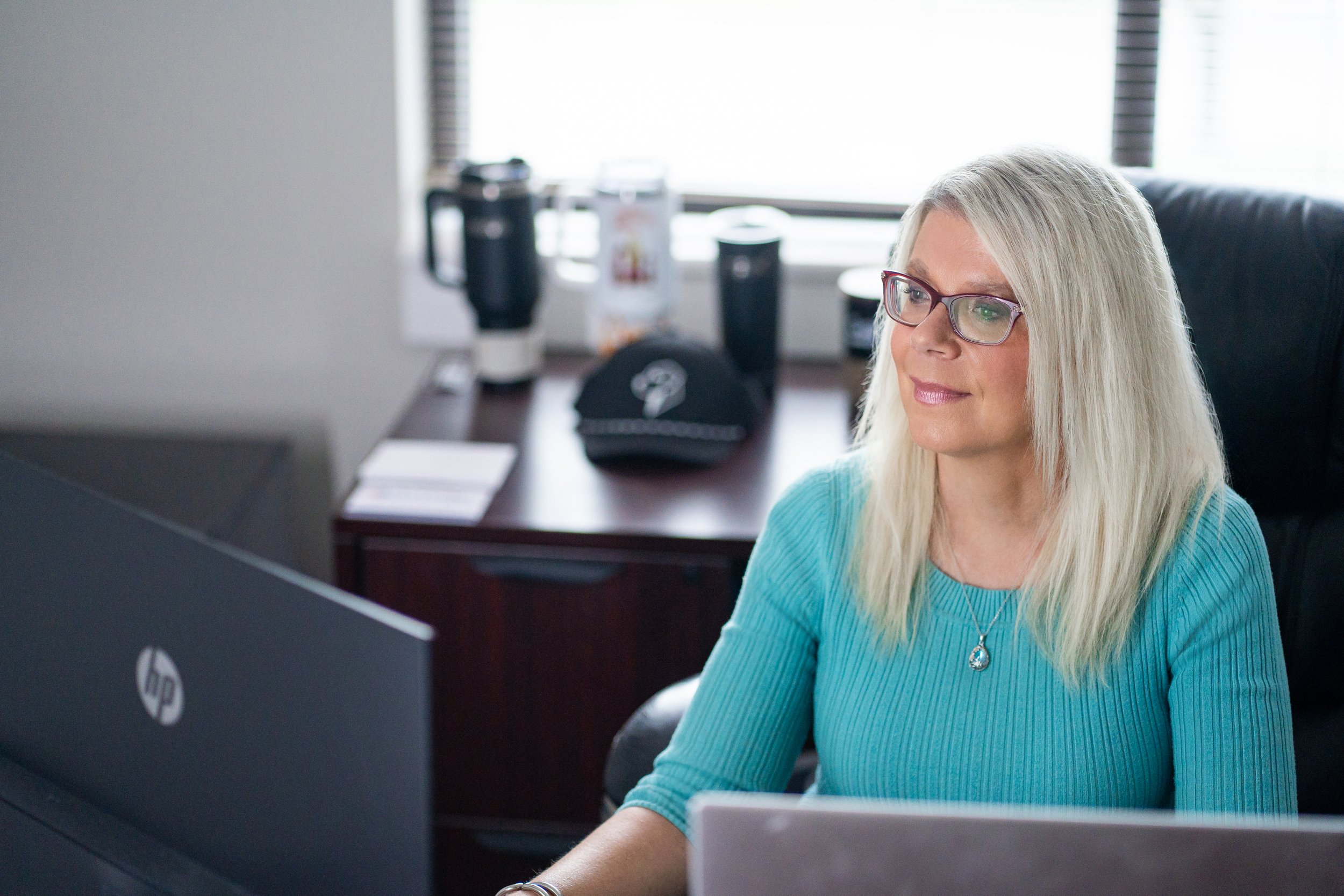A woman with long blonde hair, wearing glasses and a teal sweater, working at a desk with a computer monitor in an office.