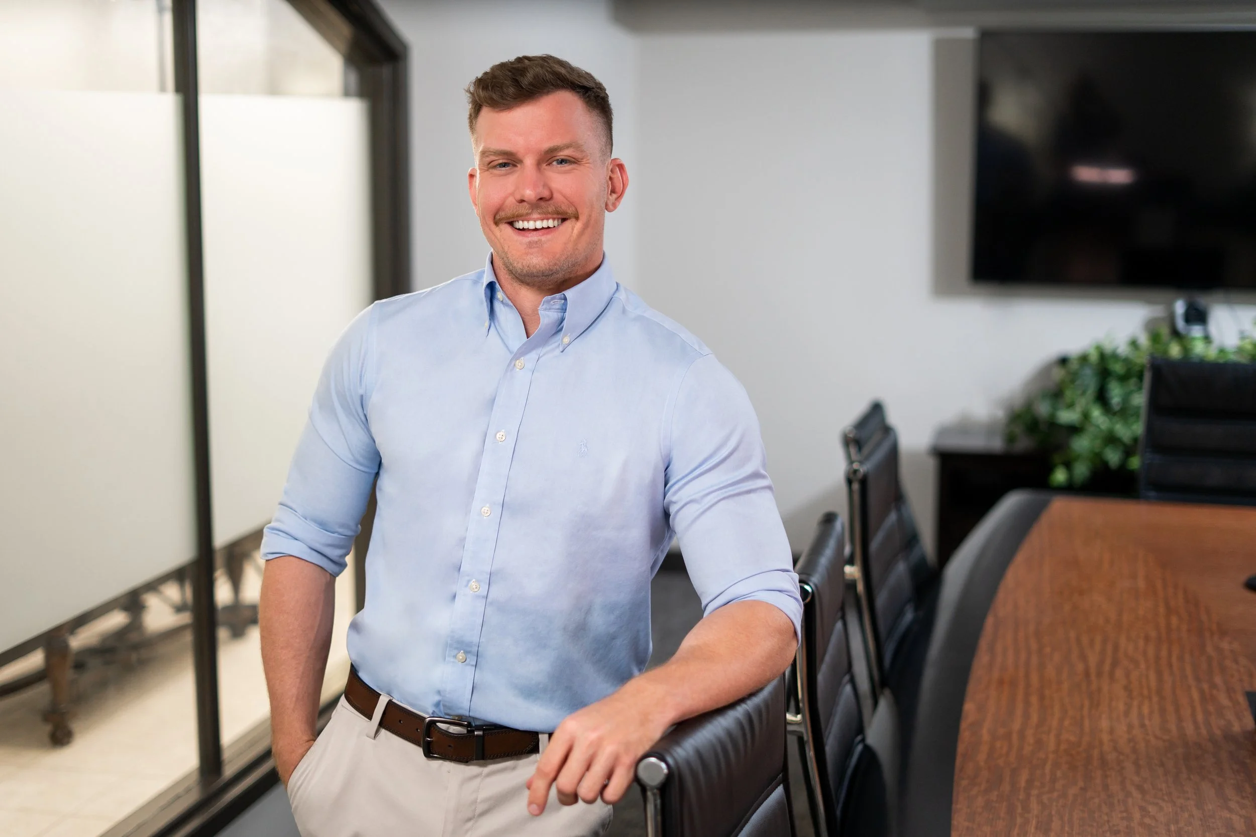 A young man with short, light brown hair, a mustache, and a beard, smiling while standing in a conference room. He is wearing a light blue button-up shirt with the sleeves rolled up and beige pants, with his right hand resting on the table. The room has a large table, chairs, some greenery, and a flat-screen TV mounted on the wall.