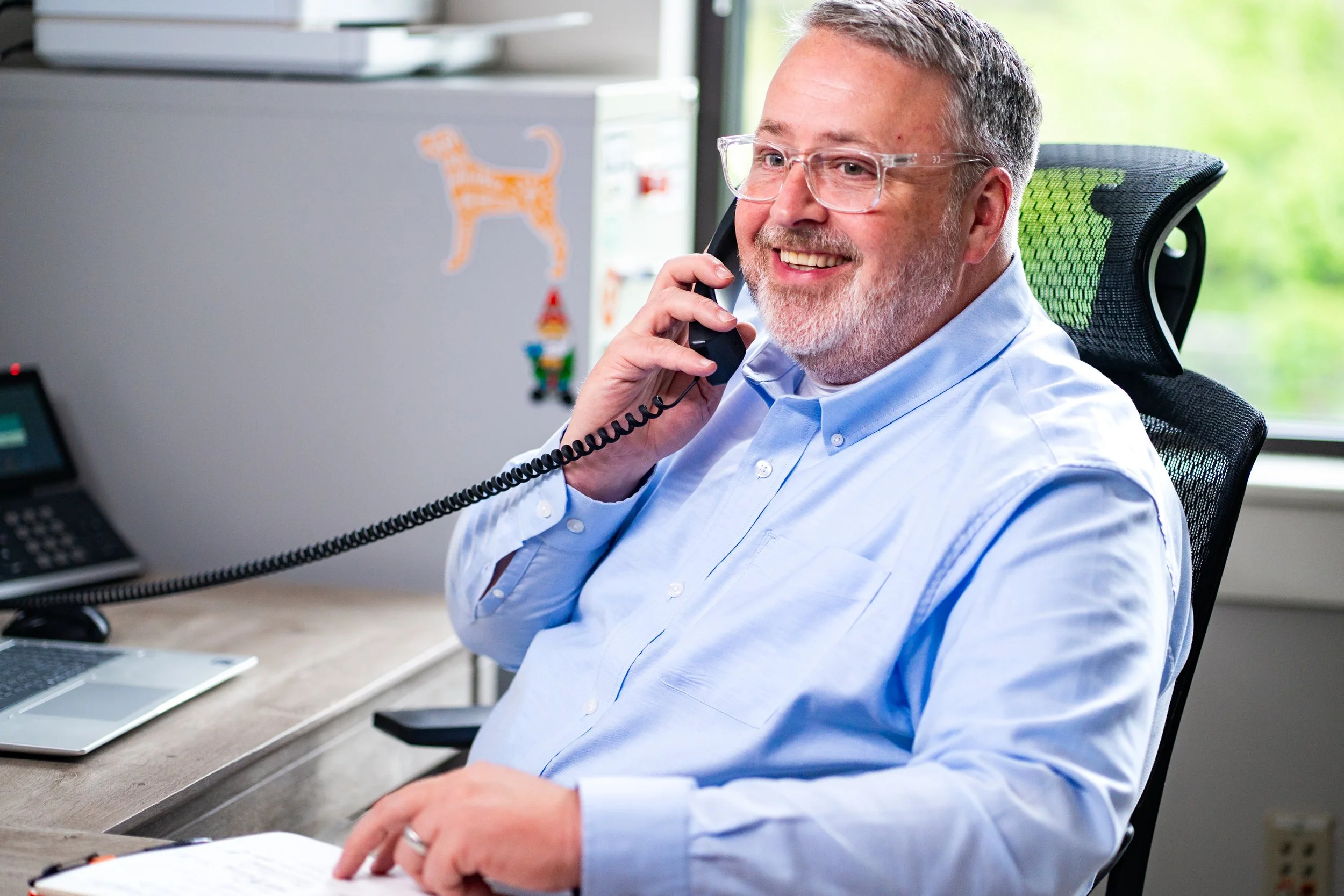 A smiling man with glasses and a beard, wearing a light blue shirt, sitting at a desk, talking on a landline phone in an office with a computer and papers.