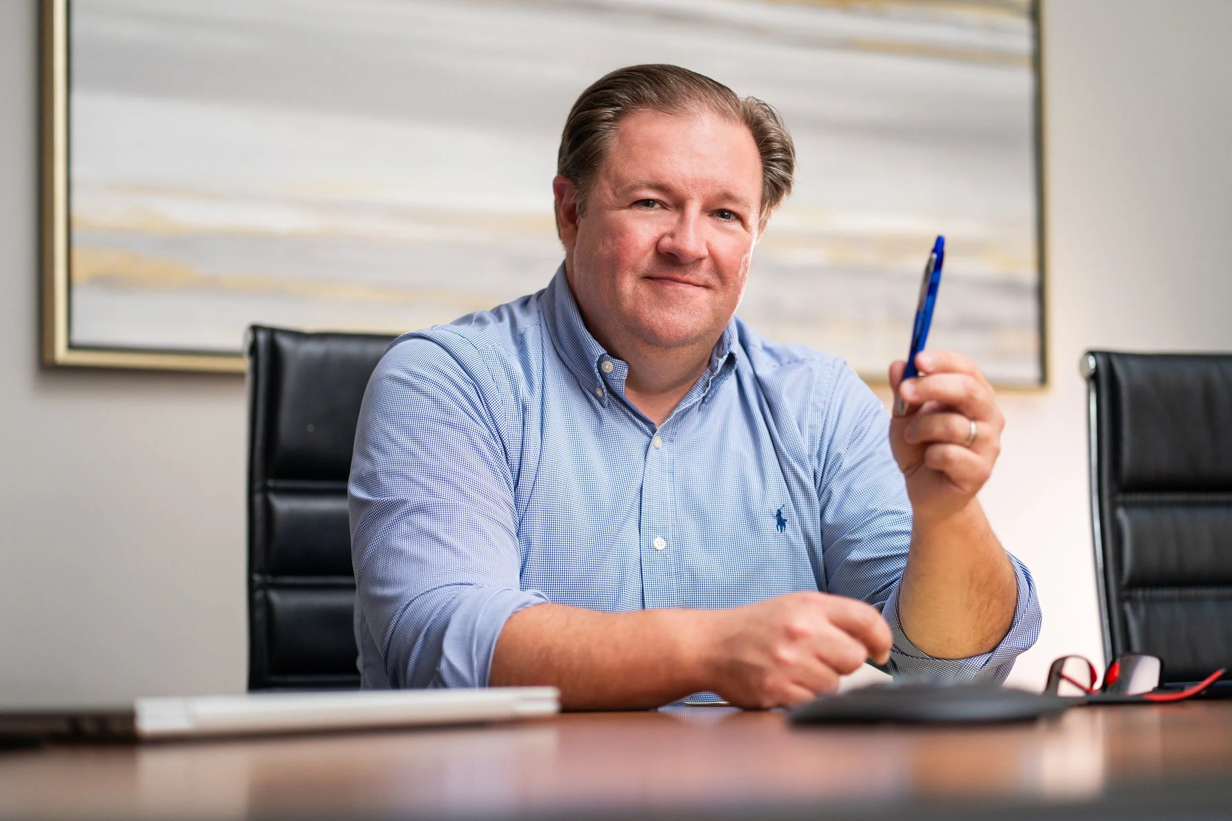 A middle-aged man with light brown hair, wearing a light blue button-up shirt, sitting at a conference table in an office. He is holding a blue pen in his right hand and looking at the camera with a slight smile. There is a large abstract painting on the wall behind him, and the table has some office supplies on it.