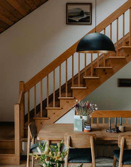 Wooden staircase with railing, hanging black pendant lamp, framed landscape painting, wooden dining table with flowers and candles, chairs with black cushions, part of a wooden ceiling and a wall with framed pictures.