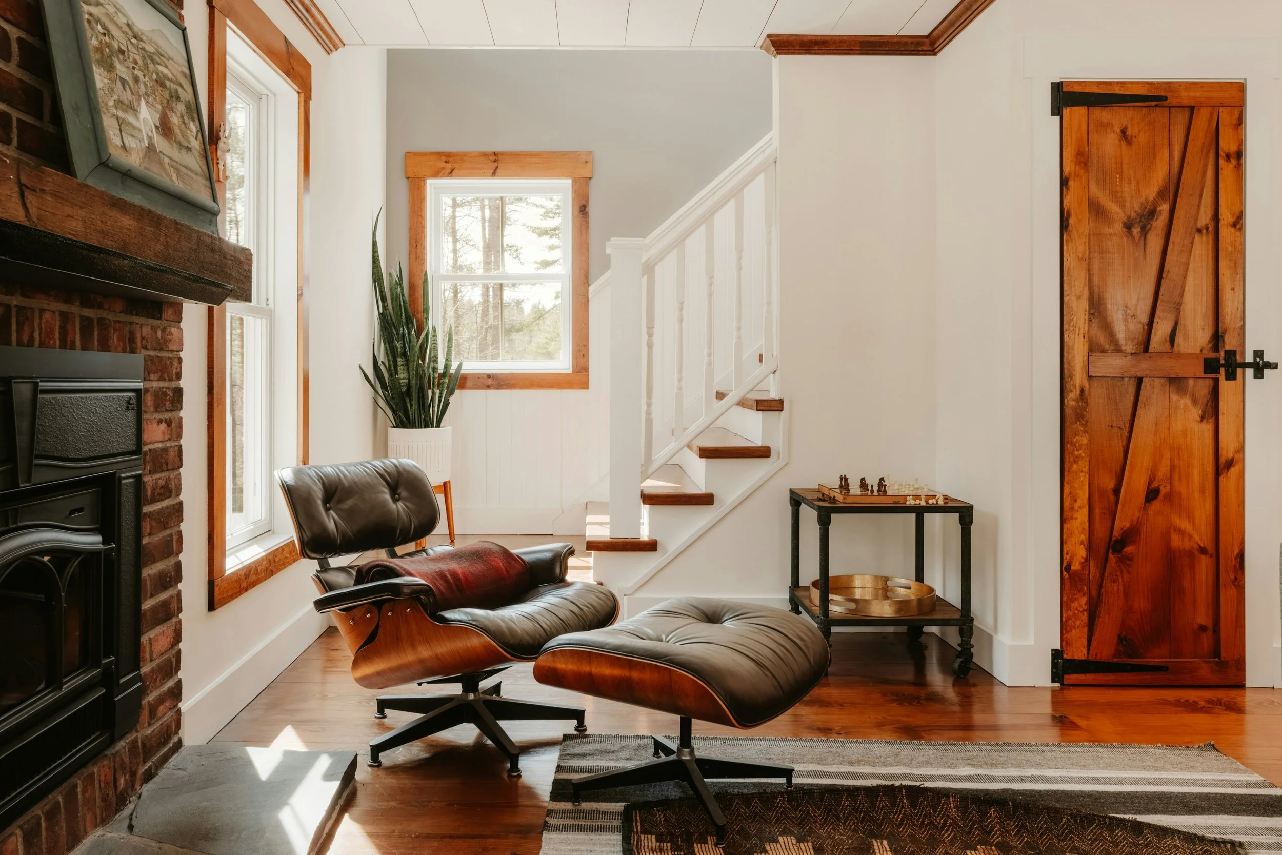 Cozy living room with a sliding barn door, a fireplace with a framed picture on the mantel, a window with wooden trim, a large potted plant, a black leather lounge chair with wooden accents, a small black side table with chess pieces, a woven basket, and a rug on a polished wooden floor.