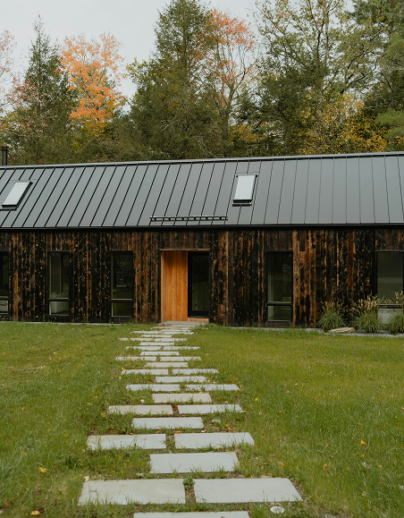 Front view of a modern house with a metal roof and dark wooden exterior, a stone pathway leading to the front door, and trees with autumn foliage in the background.