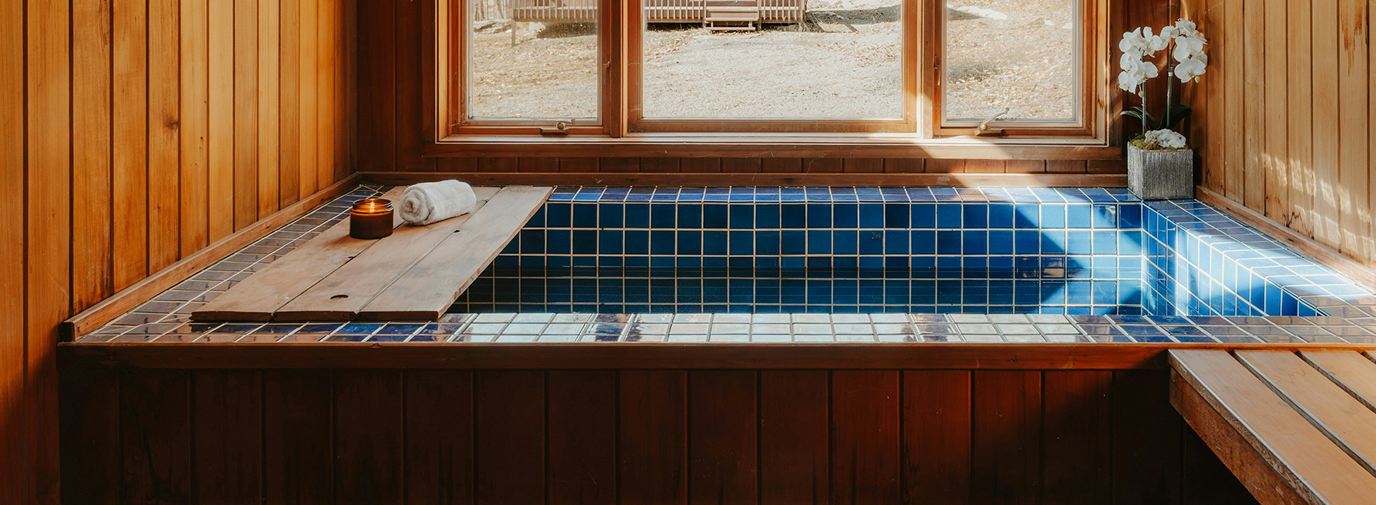 Indoor wooden hot tub with blue tiles, a small wooden plank over part of it, a rolled white towel, a candle, and a potted plant with white flowers, near a large window